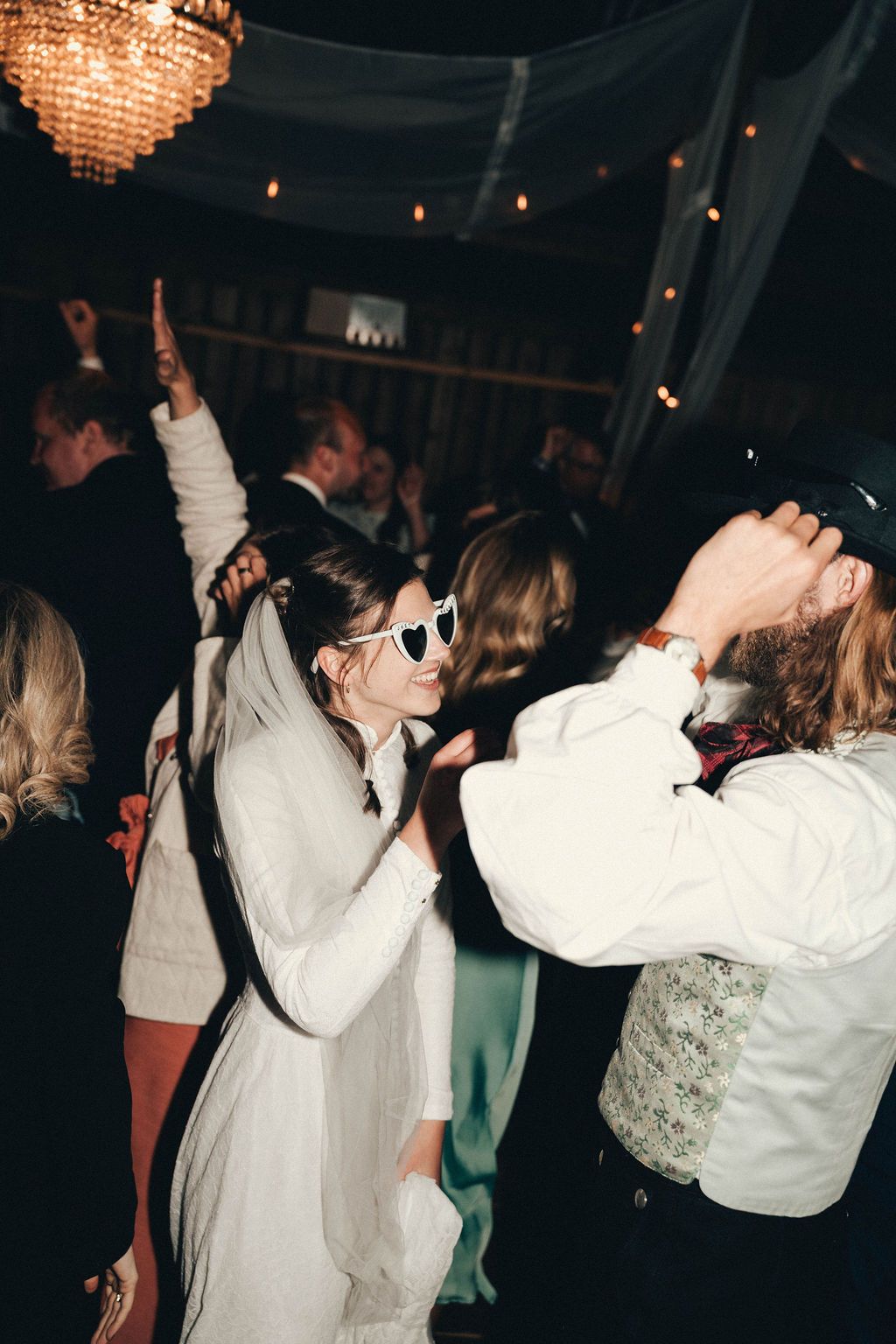 People dancing and celebrating under crystal chandeliers and string lights at an indoor party venue.