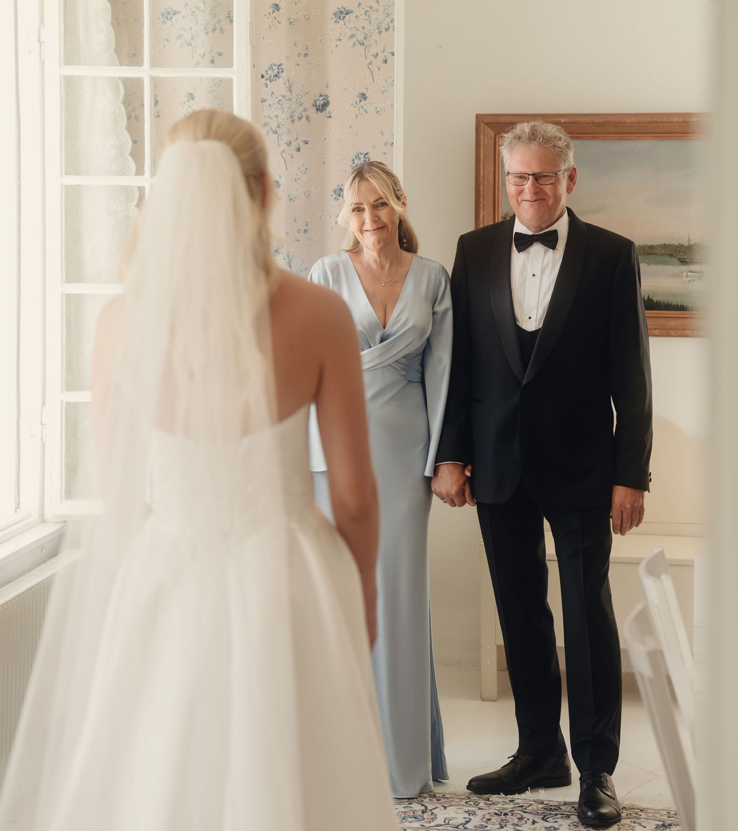 Bride in a white gown and veil faces her smiling parents, a man in a tuxedo and a woman in a blue gown, in a bright room.