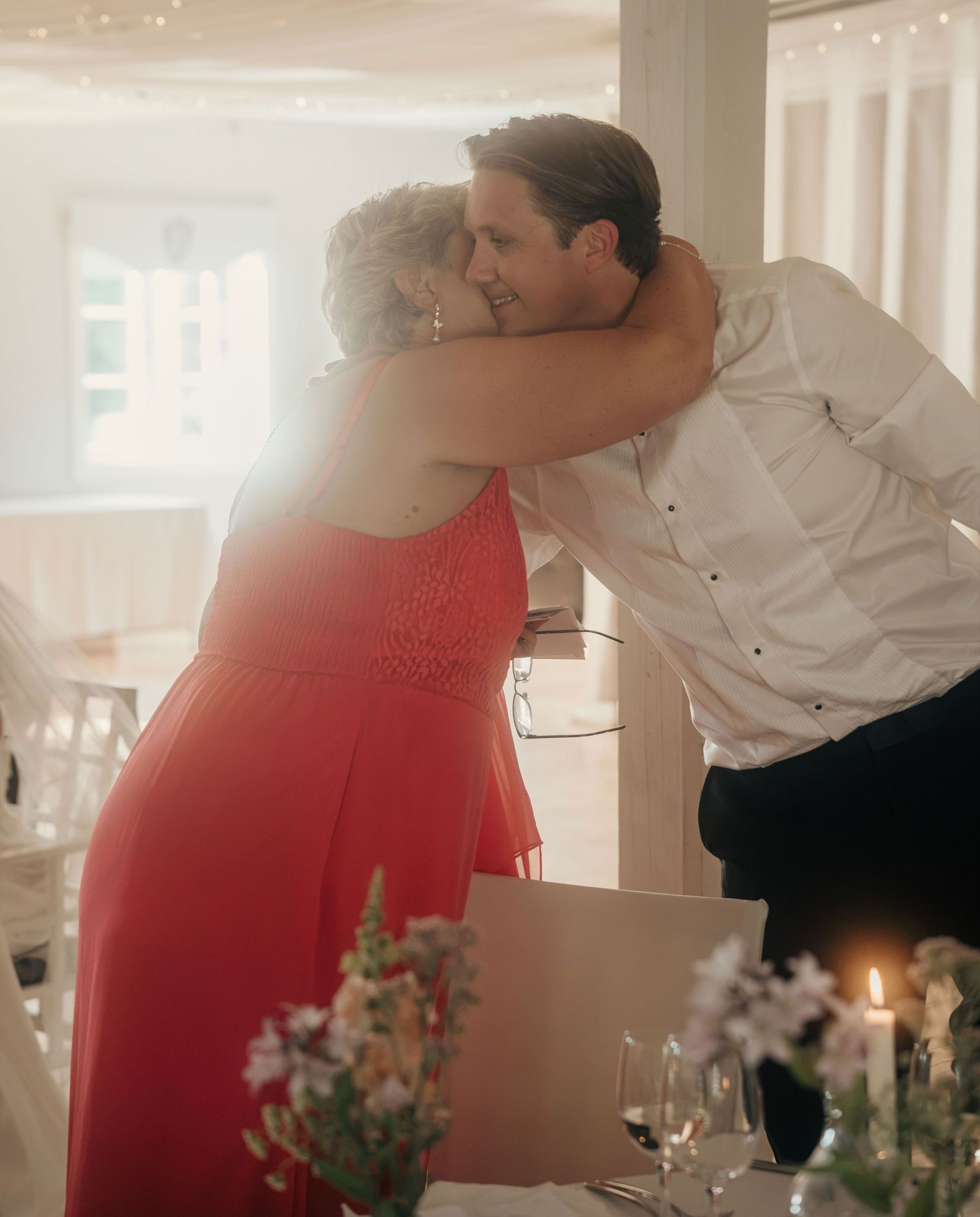 Man in a white shirt hugs a woman in a coral dress at a candlelit wedding reception with flowers on the table.