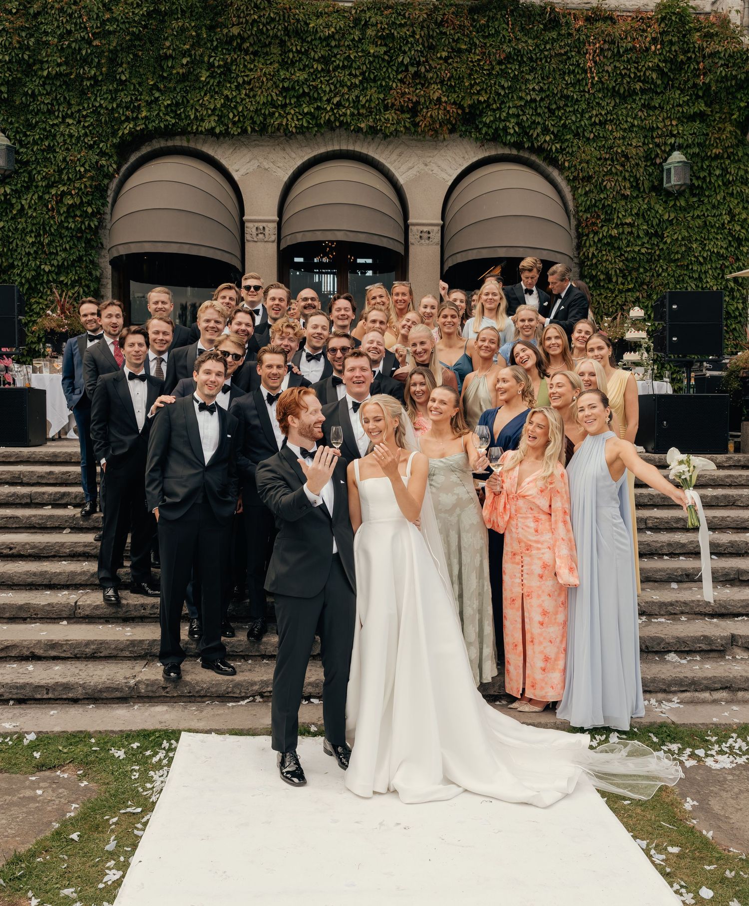 Bride and groom celebrate on a white carpet in front of wedding guests gathered on stone steps by an ivy-covered building.