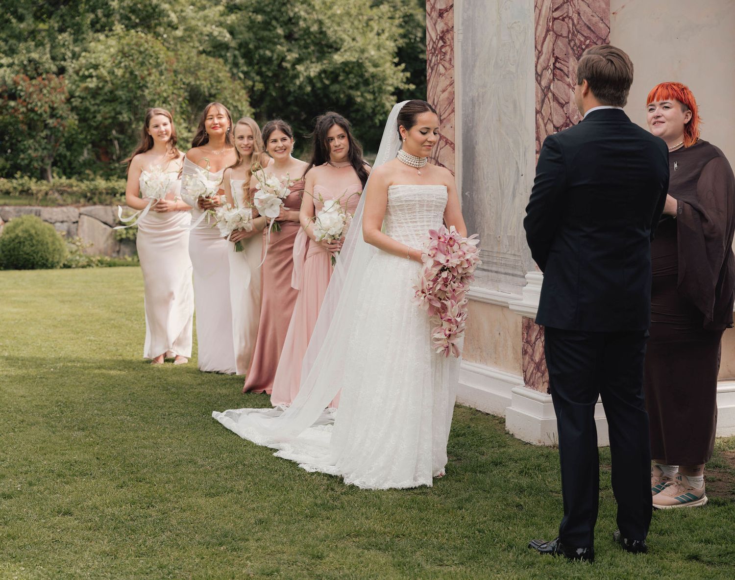 Bride in white lace gown with bridesmaids in pink and ivory dresses holding bouquets at outdoor wedding ceremony.