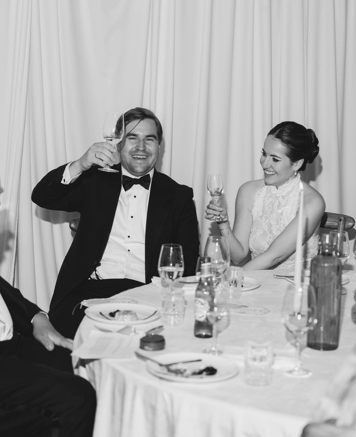 Black and white photo of a couple in formal attire toasting with champagne glasses at a wedding reception dinner.