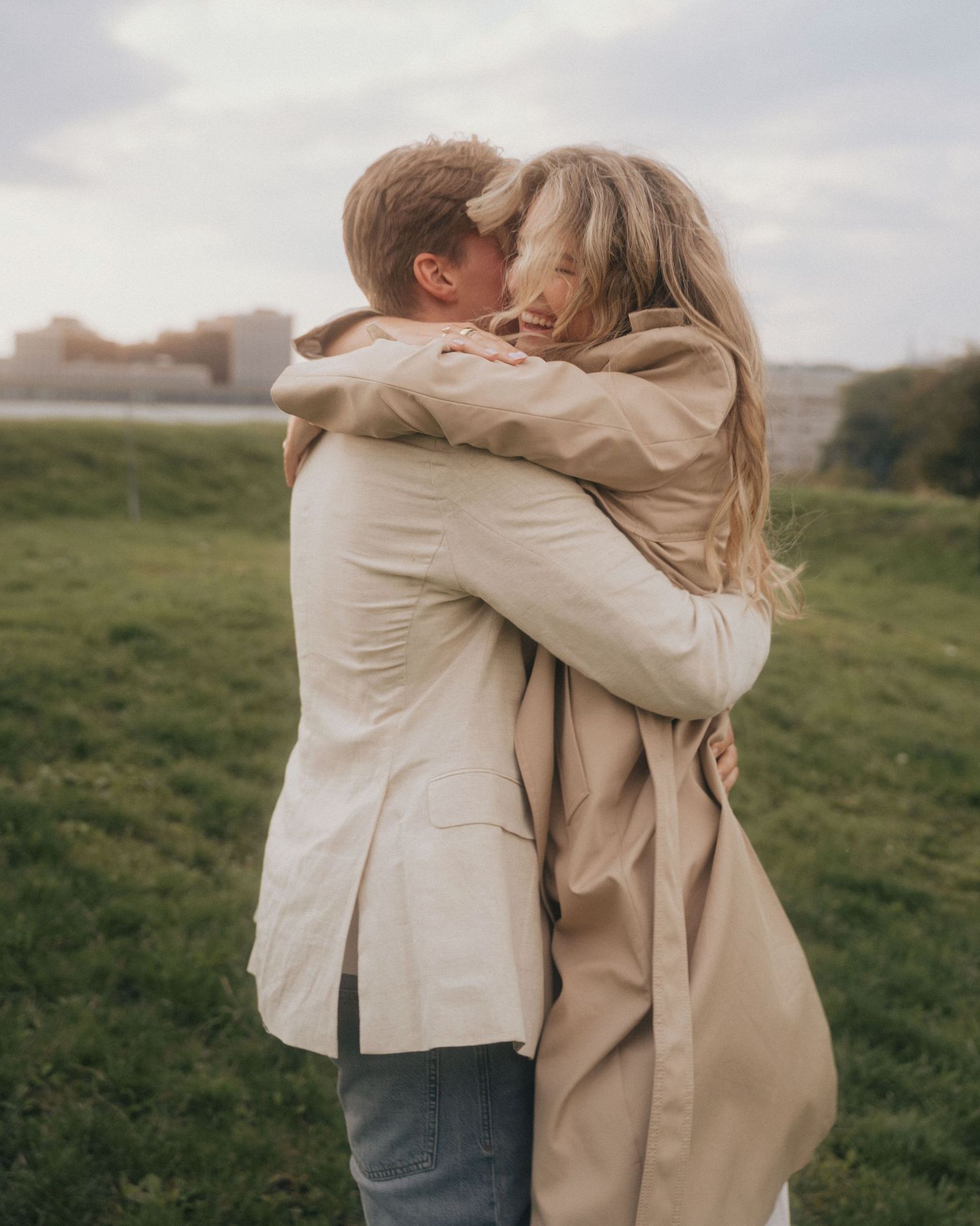 Two people embrace tenderly in a field during sunset, wearing neutral-toned clothing in a romantic outdoor setting.