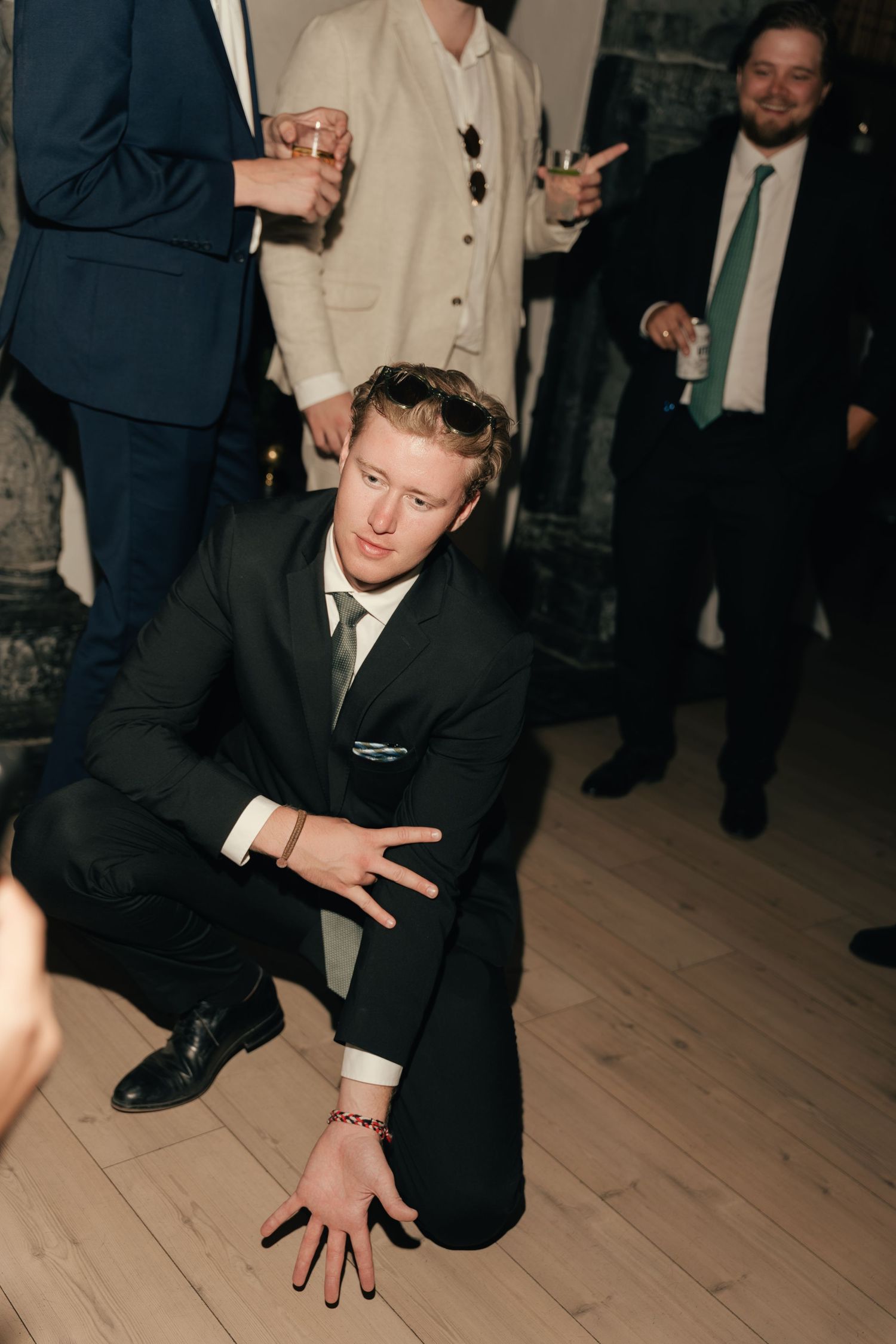 Wedding guest in black suit crouches on dance floor during lively indoor wedding reception.