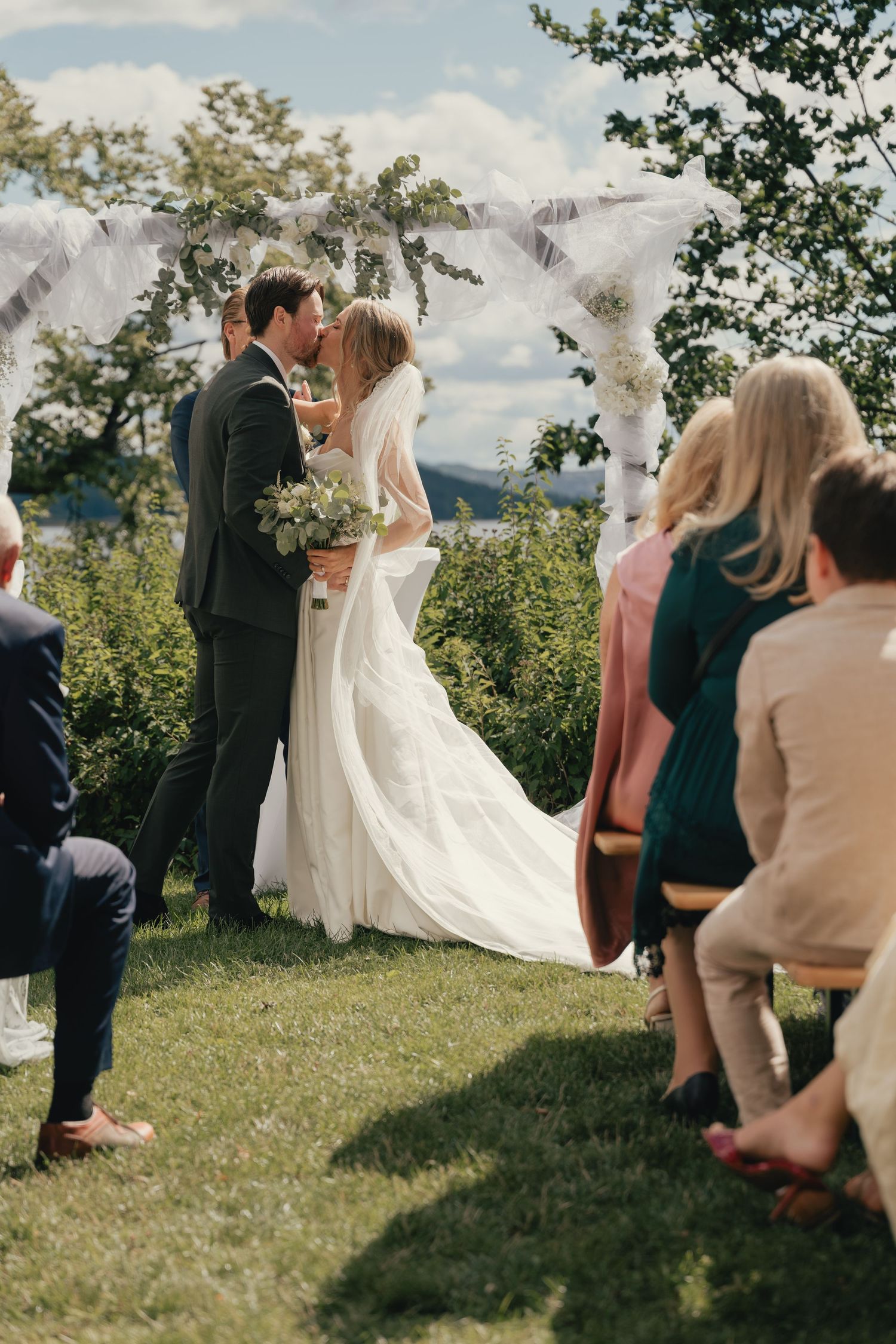 Bride and groom share first kiss at outdoor wedding ceremony beneath floral arch with guests watching.