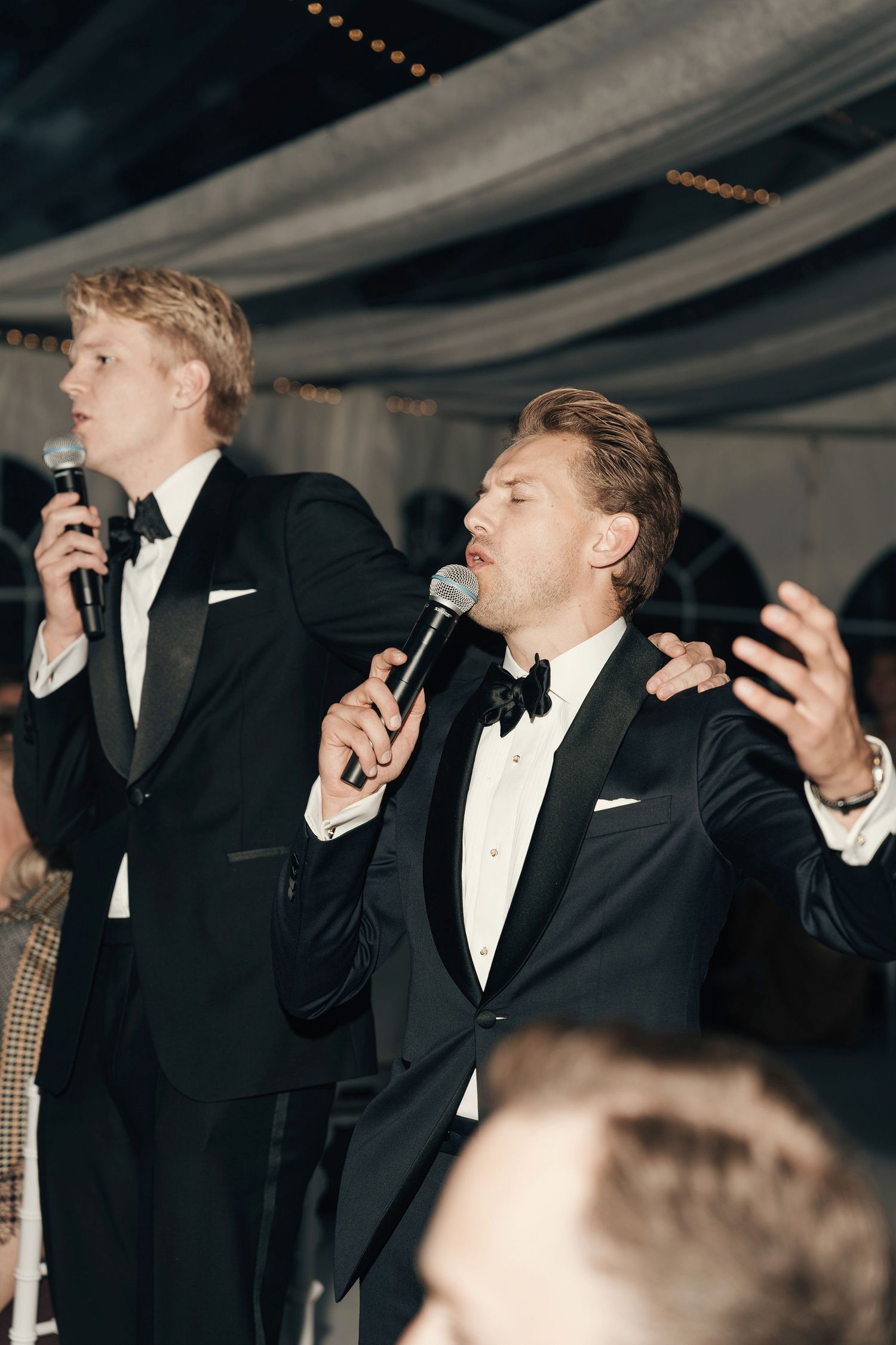 Two men in black tuxedos sing into microphones and gesture energetically during a wedding reception.