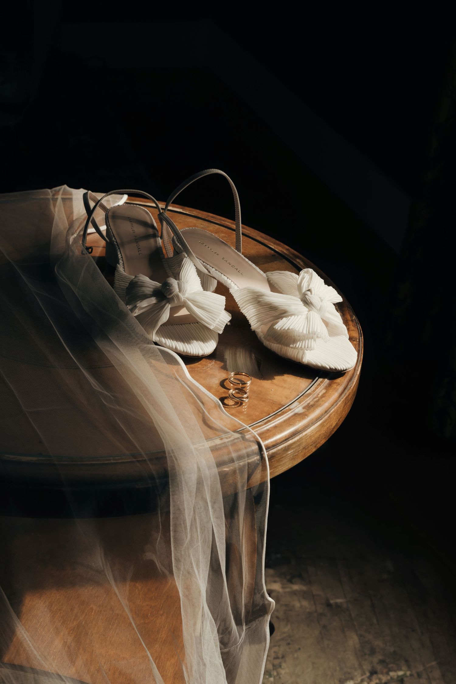 White satin bow wedding sandals and two gold rings rest on a round wooden table beneath a sheer bridal veil in dramatic, moody light.