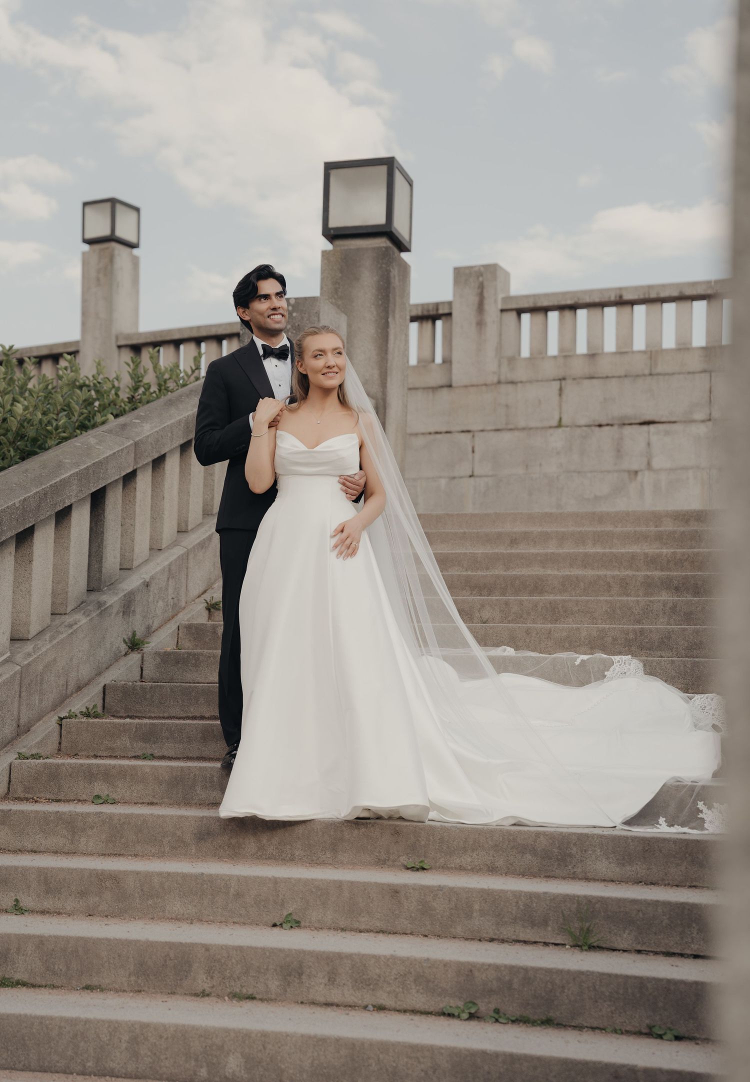 Bride in a flowing white gown and long veil stands with her groom in a tuxedo on grand stone steps at their wedding.