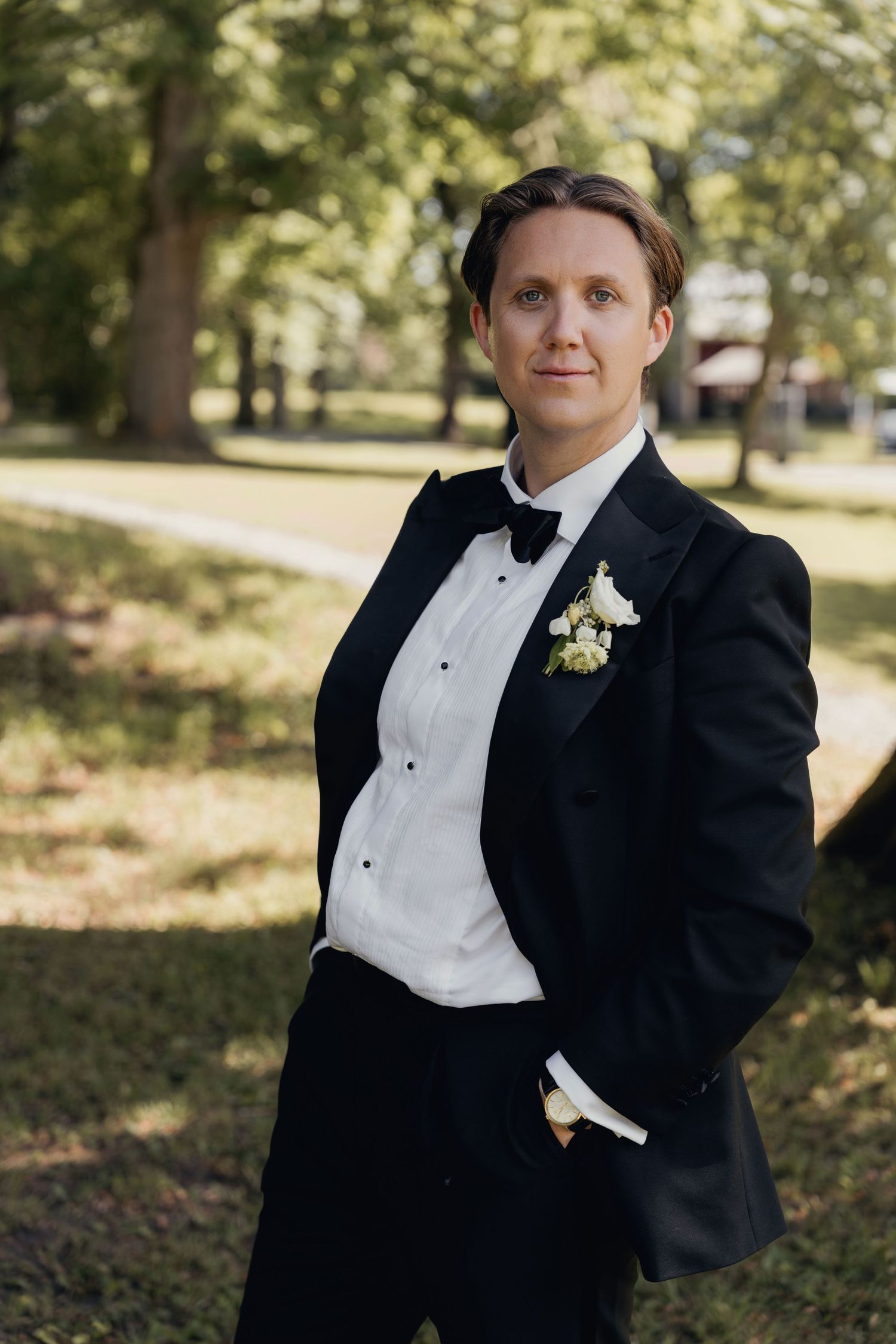 A groom in a black tuxedo with a bow tie and white floral boutonniere stands with a hand in his pocket in a sunlit park.