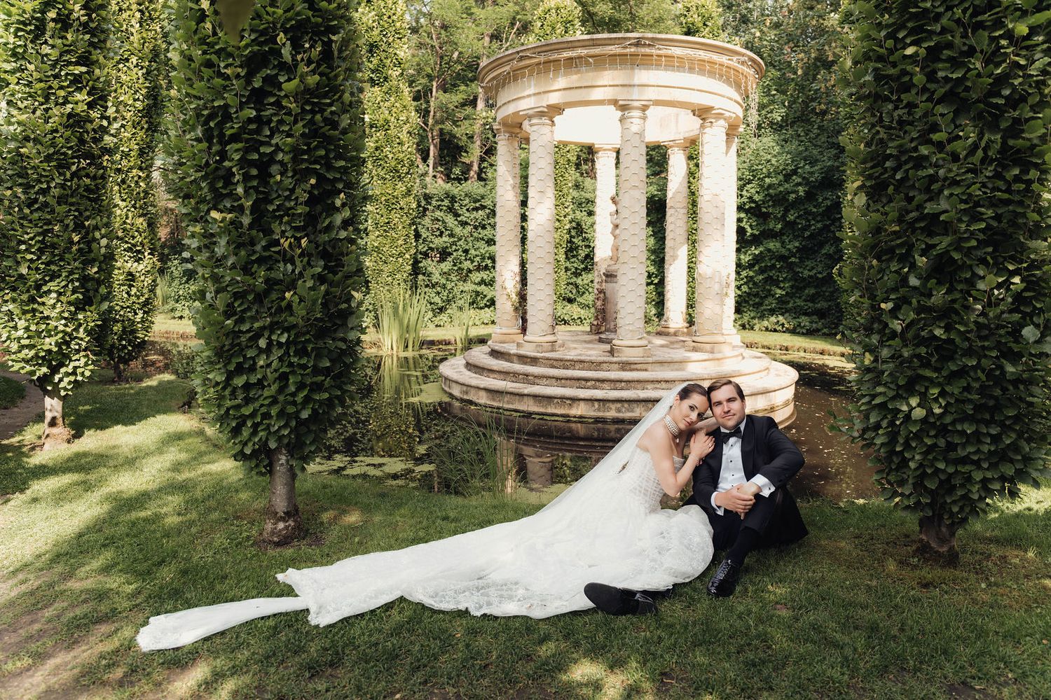 Bride and groom sitting together on grass in front of a stone rotunda with columns surrounded by lush greenery.