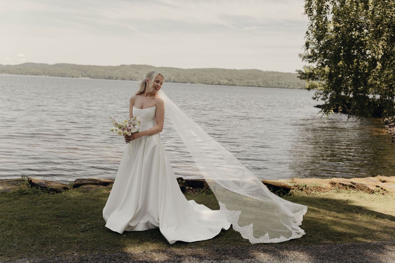 Bride in an elegant white gown and long flowing veil holds a bouquet while standing on a grassy lakeside shore on her wedding day.