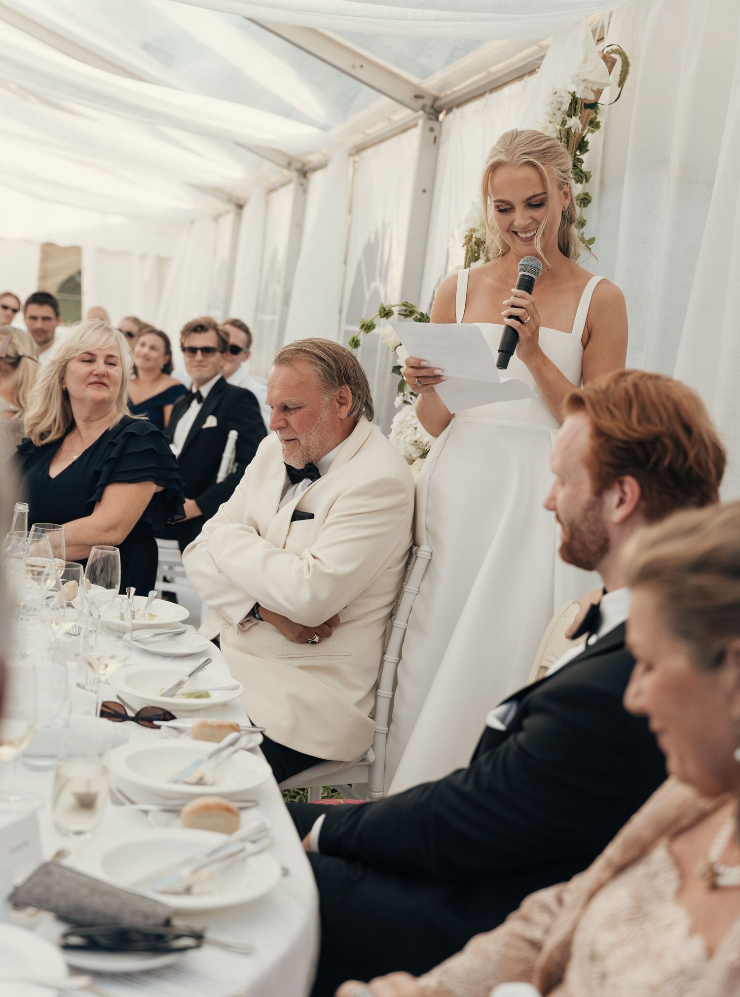 Bride speaks into a microphone during a wedding reception toast while the groom and seated guests look on.
