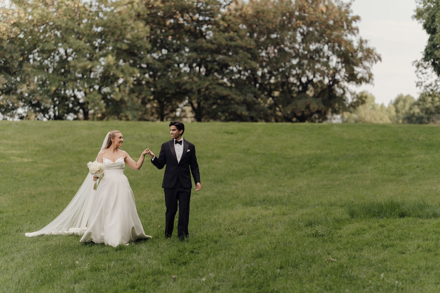 Bride and groom holding hands walking on lush green grass with trees in background on their wedding day.