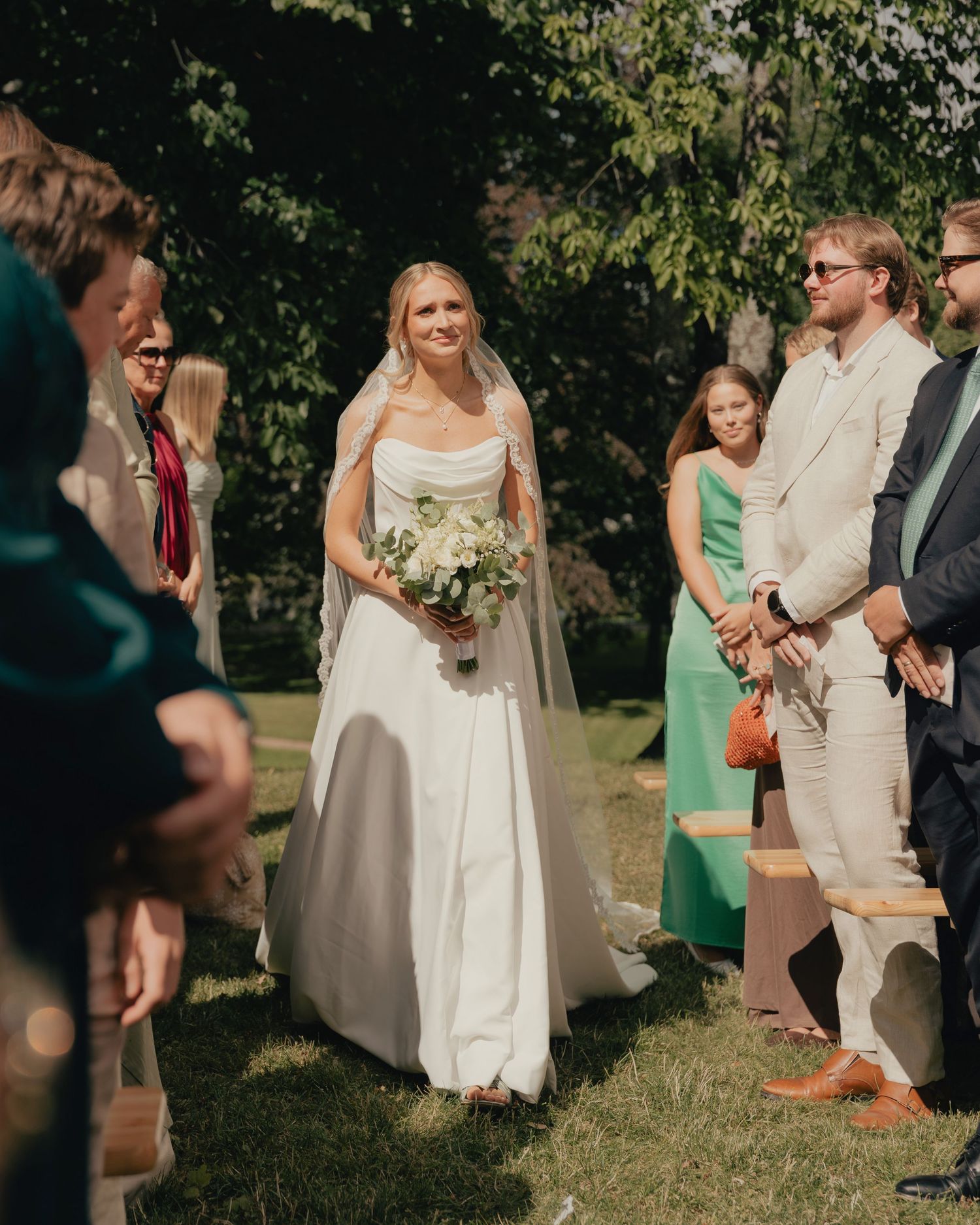 Bride in white gown carrying bouquet walks down outdoor aisle surrounded by wedding guests.