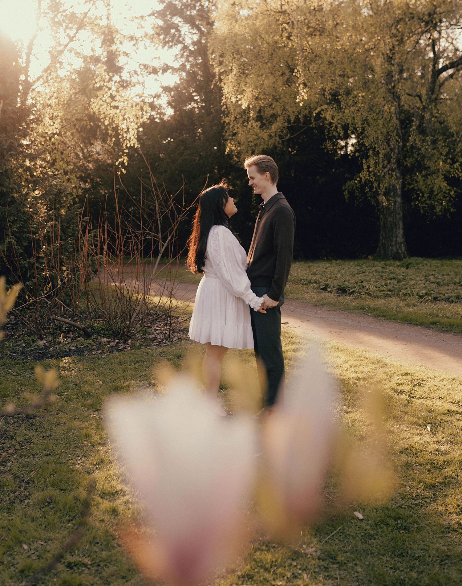 A romantic couple stands together on a path at sunset with soft bokeh flowers in the foreground.