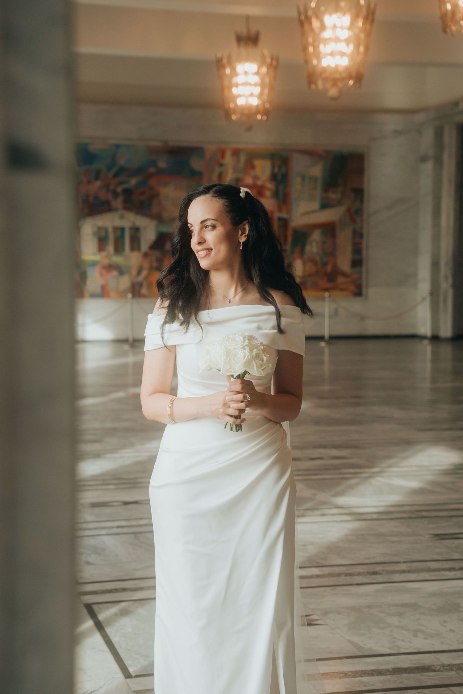 A person in a white wedding dress stands in an elegant venue with crystal chandeliers overhead.