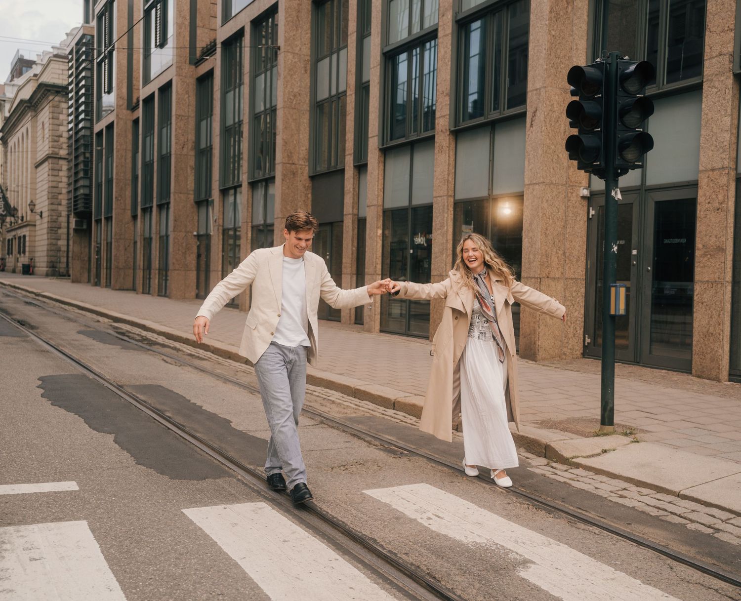 Two people in light colored outfits walk down a narrow cobblestone street between tall buildings.