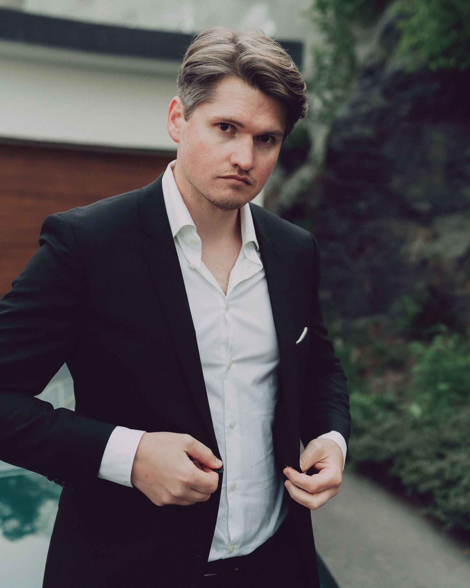 A person in a black blazer and white shirt adjusts their collar against a blurred outdoor background.
