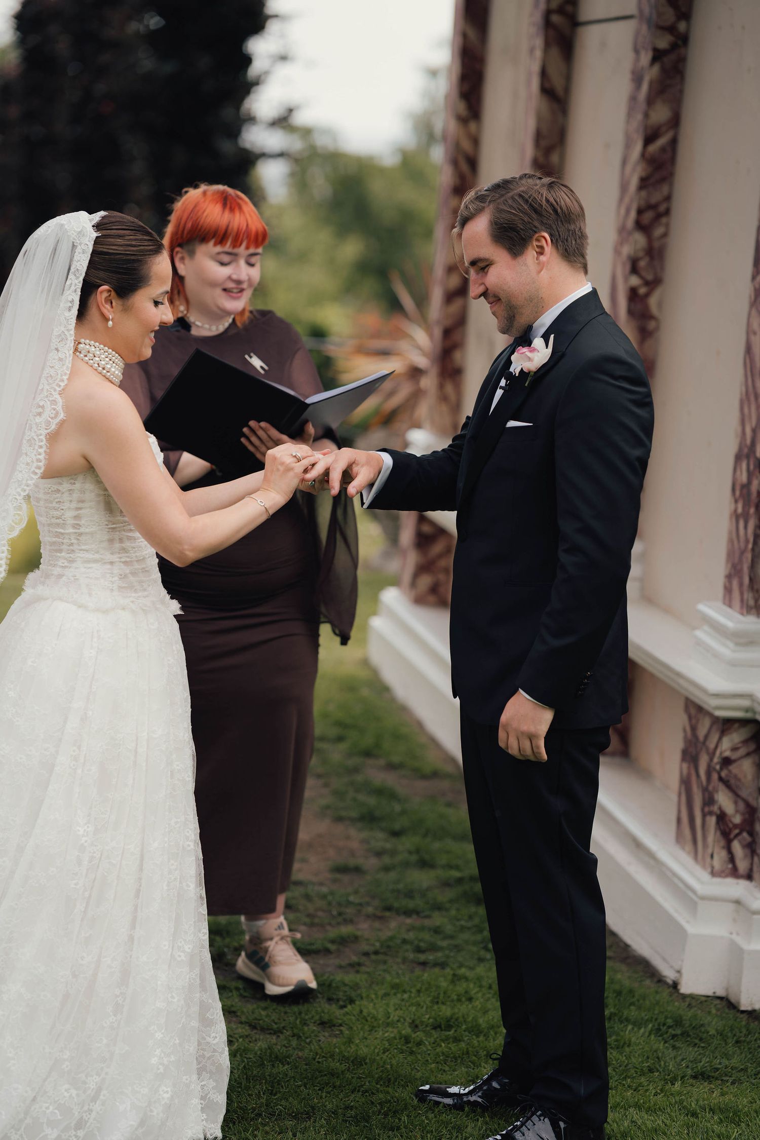 Bride placing wedding ring on groom's finger during outdoor ceremony, officiant with red hair holding book behind them.