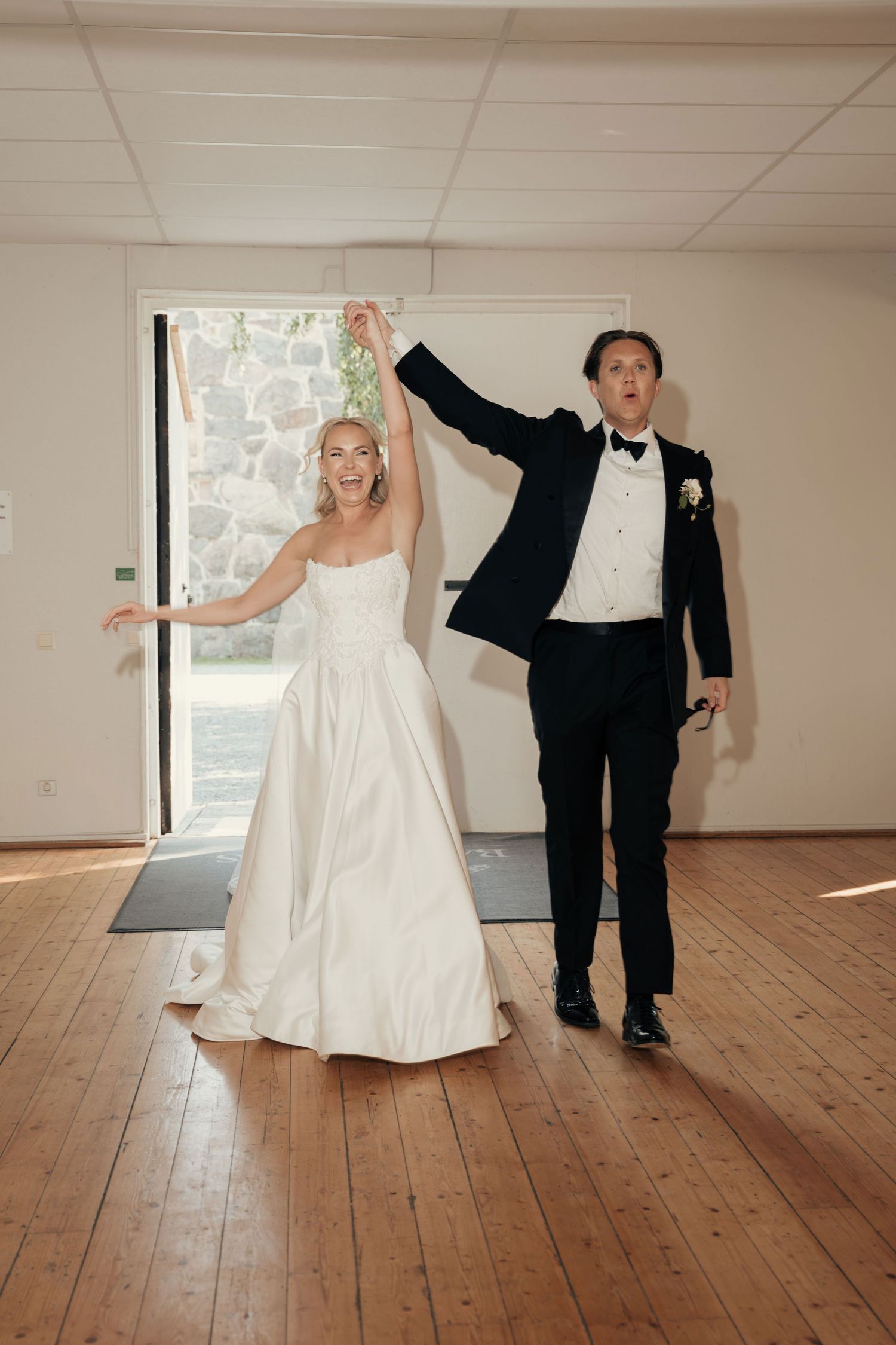 Bride in a strapless white gown and groom in a black tuxedo raise their arms joyfully as they enter a bright wooden-floored venue.