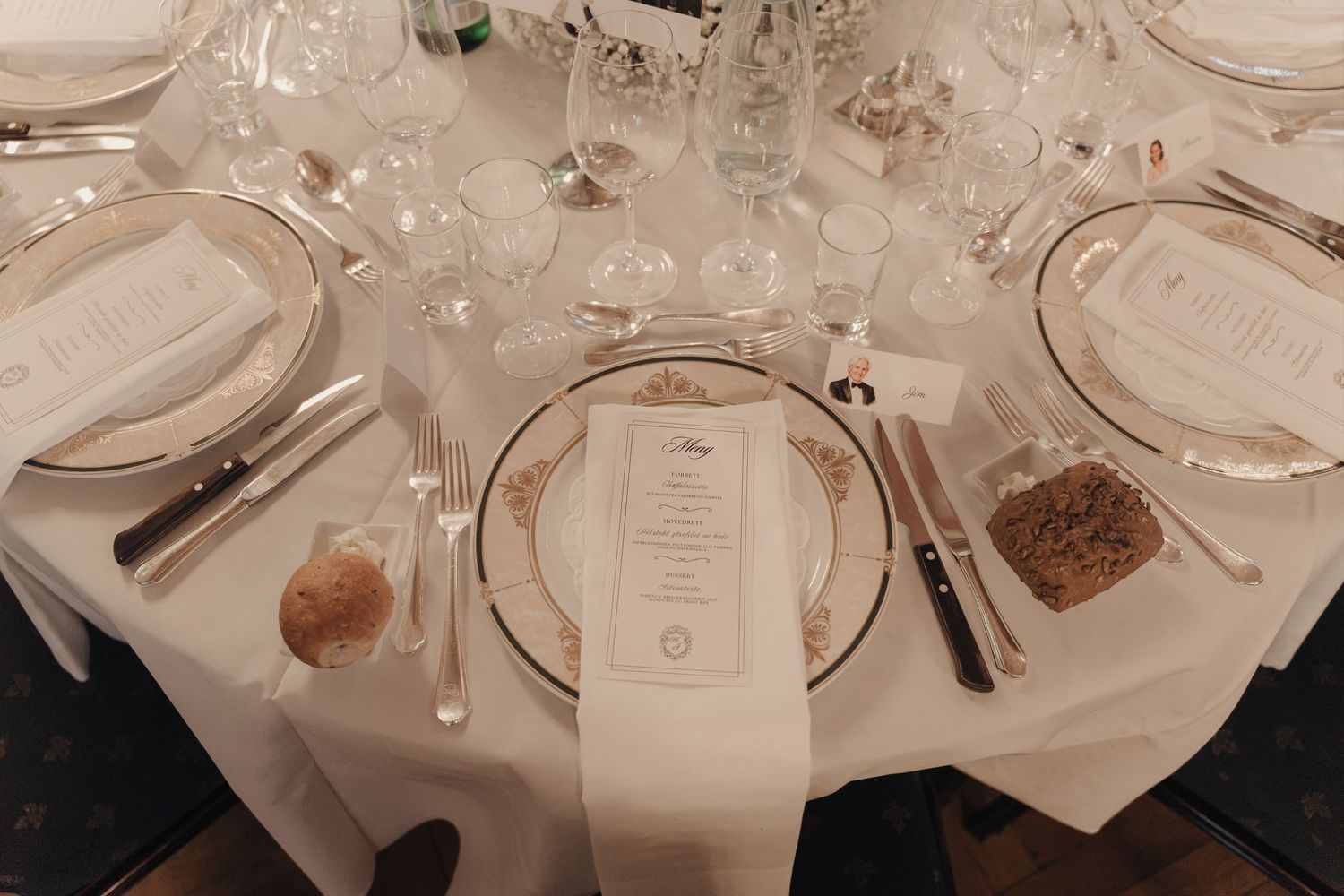 Elegant banquet table setting with a printed menu on a folded white napkin, gold-rimmed china plates, silverware, wine glasses and bread rol