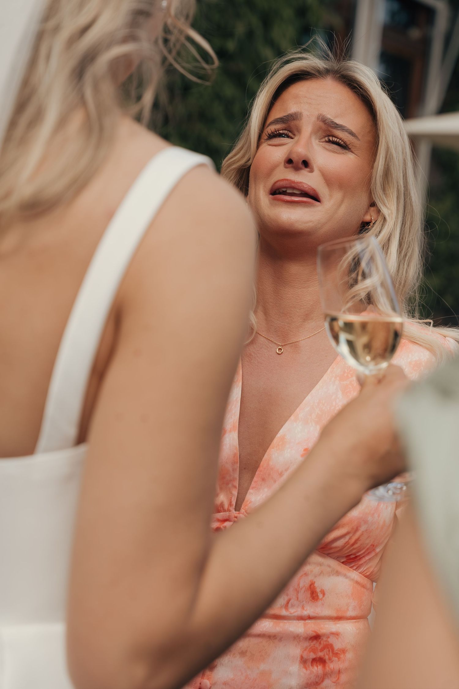 Emotional woman in a coral floral dress tears up while holding a champagne glass during a wedding celebration.