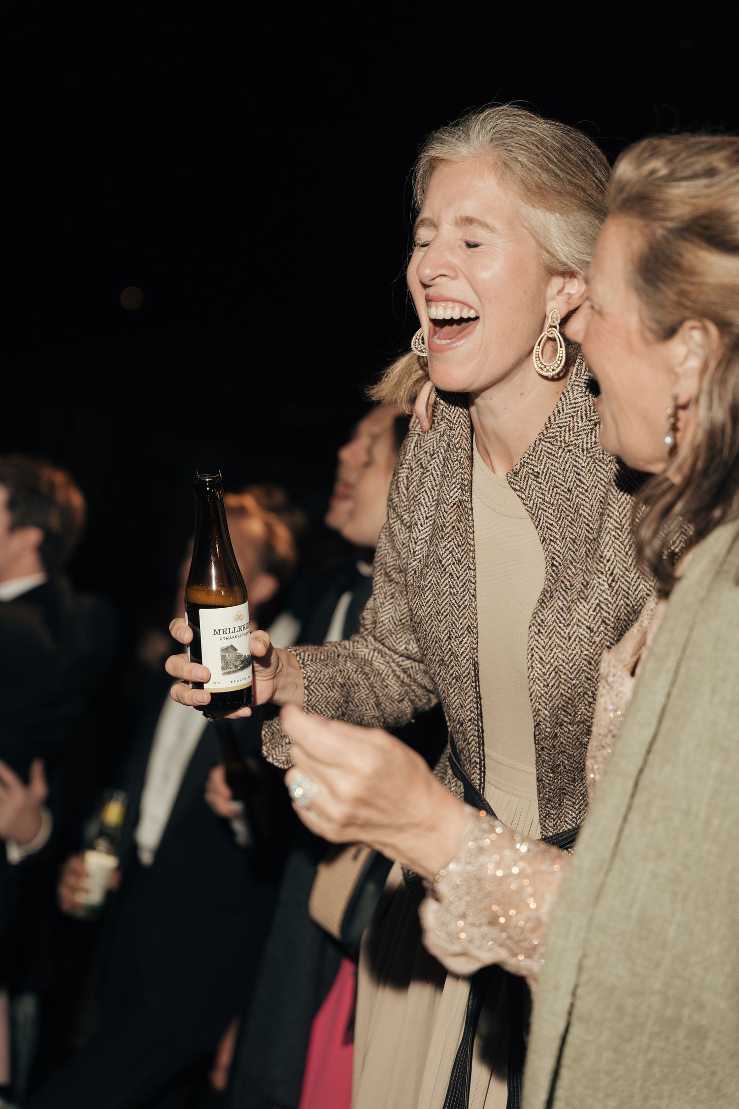 A laughing woman in a tweed blazer and drop earrings holds a beer bottle while mingling with guests at an evening wedding celebration.