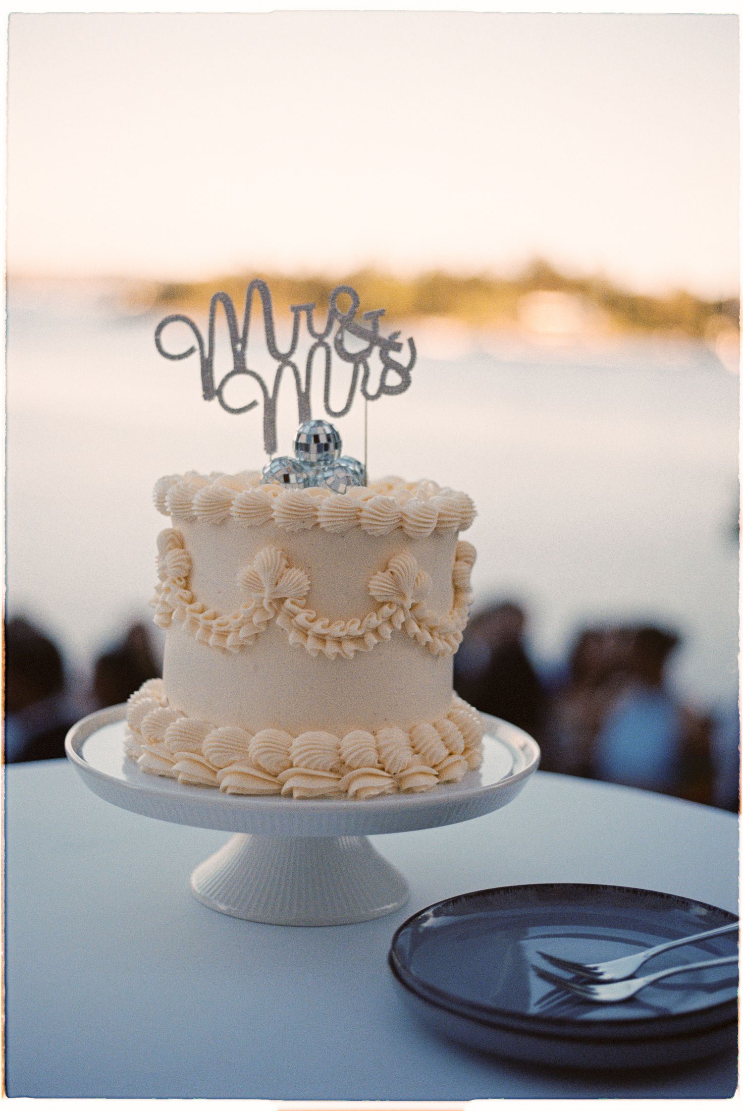 Single-tier white wedding cake with Mr and Mrs topper on pedestal at sunset.