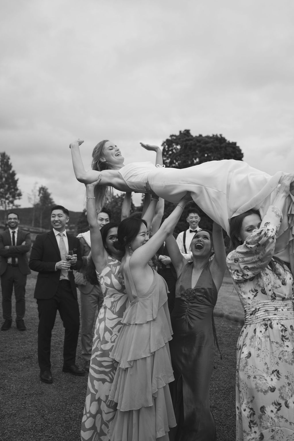 Wedding guests lift and support a person in a white dress during an outdoor celebration, captured in black and white.