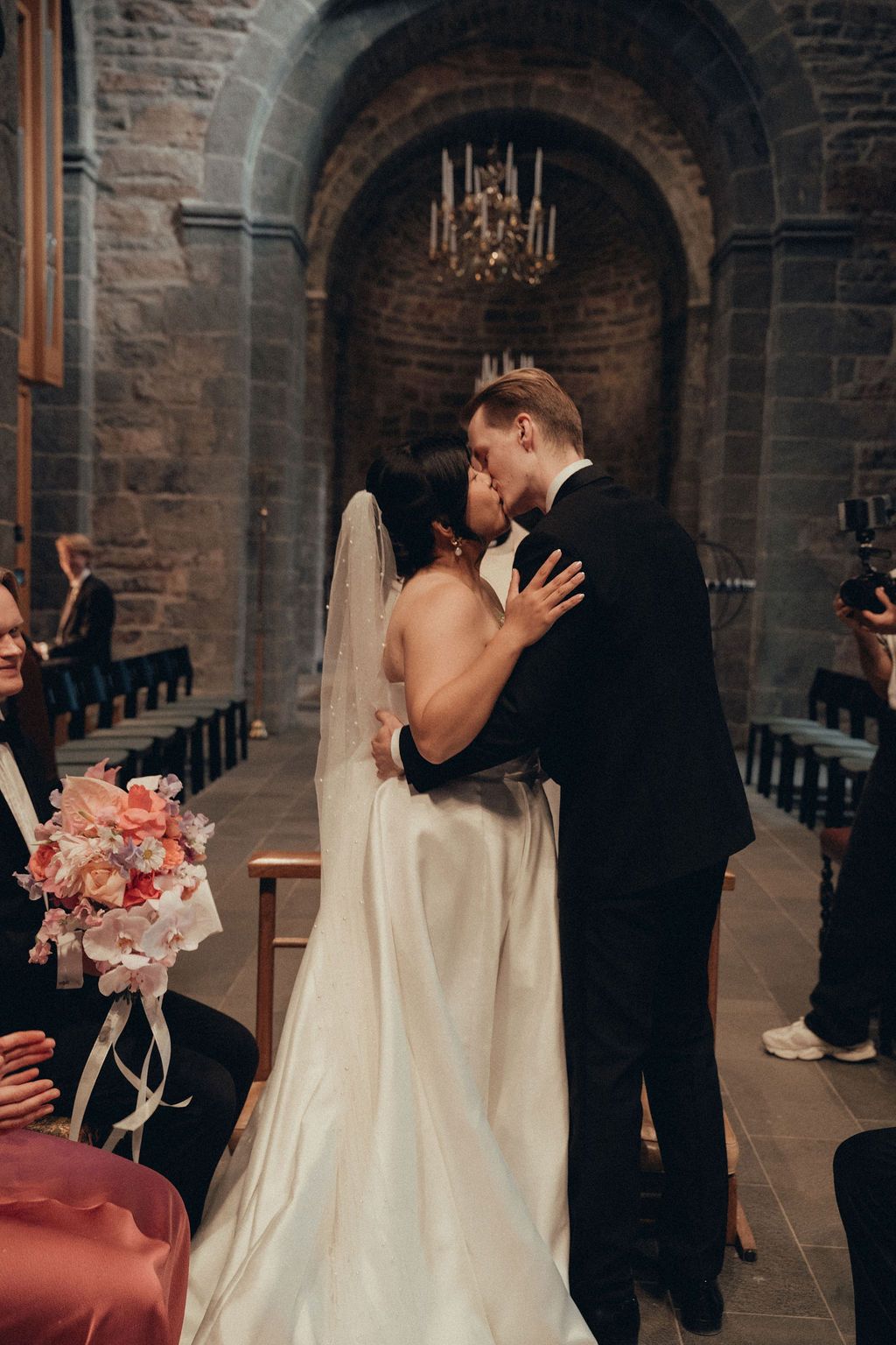 Newlyweds share their first kiss during a wedding ceremony inside a historic stone church with candlelit archways.