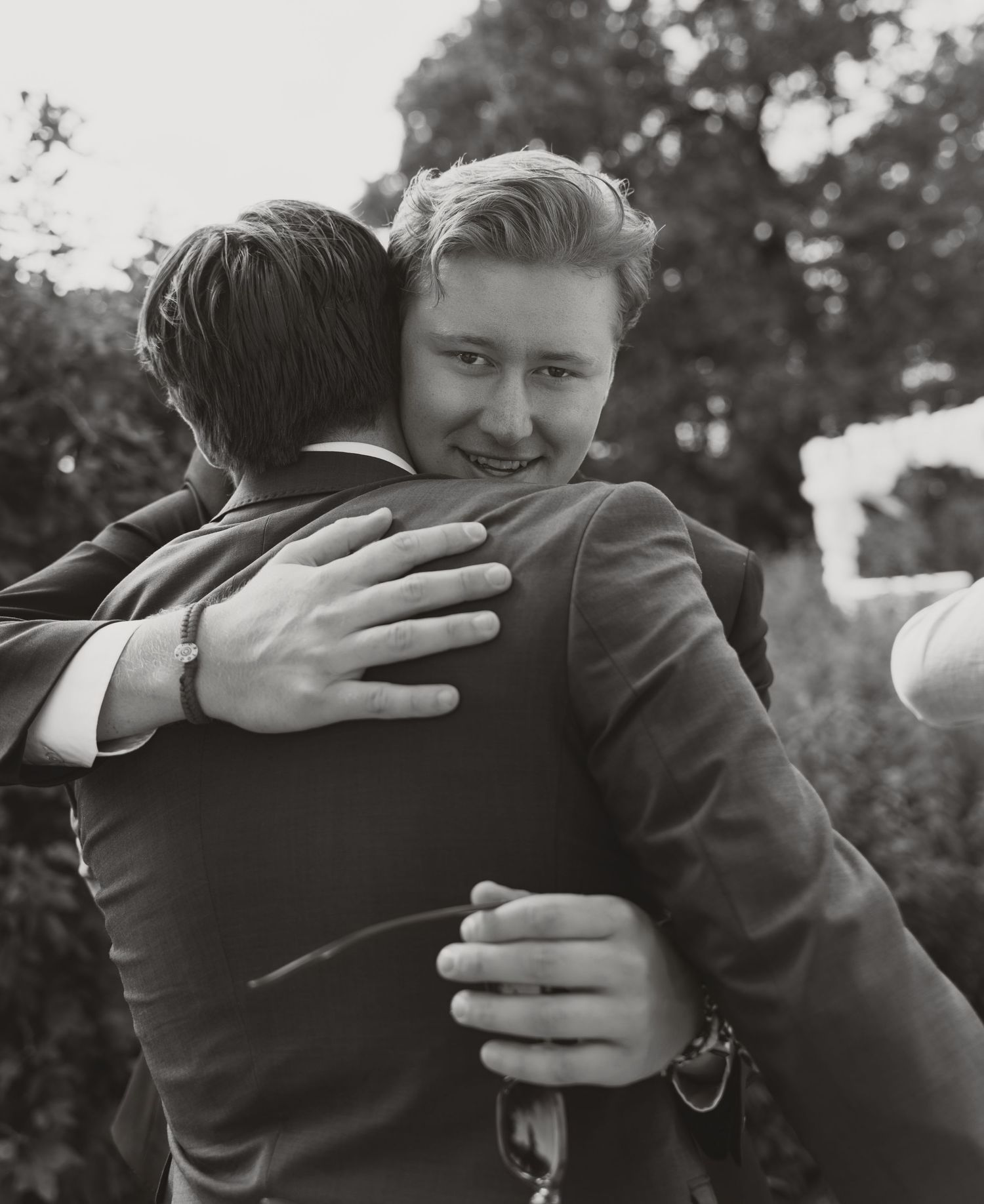 Two men in suits share a warm embrace outdoors at a wedding celebration in black and white photo.