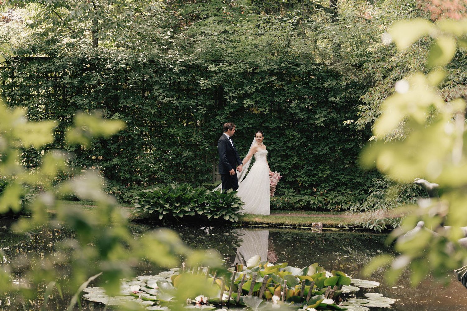 A bride and groom stand hand in hand by a tranquil pond surrounded by lush green foliage in a romantic garden setting.
