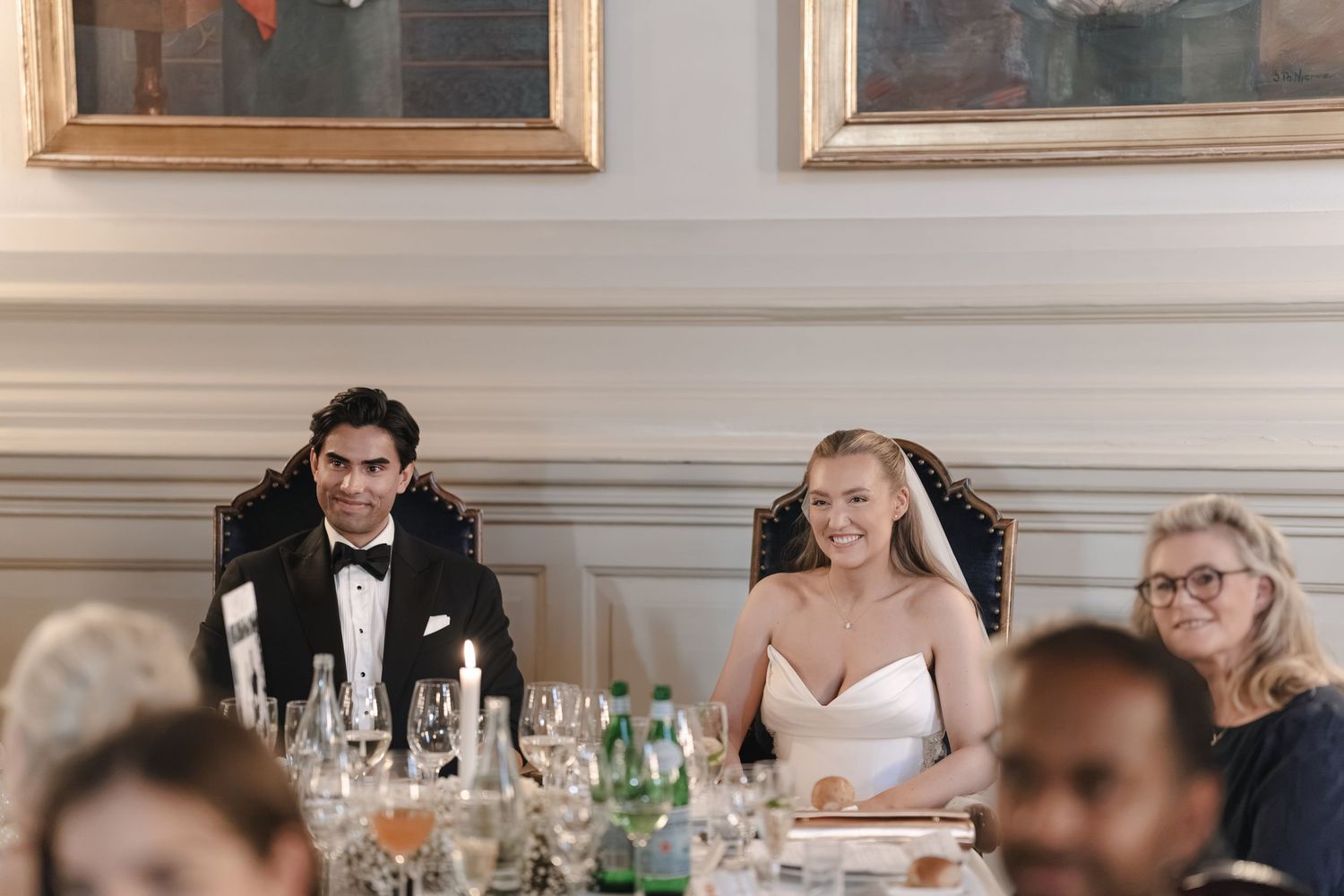A smiling bride in a white dress and a groom in a black tuxedo sit at an elegant wedding reception table with candles, glassware and guests.