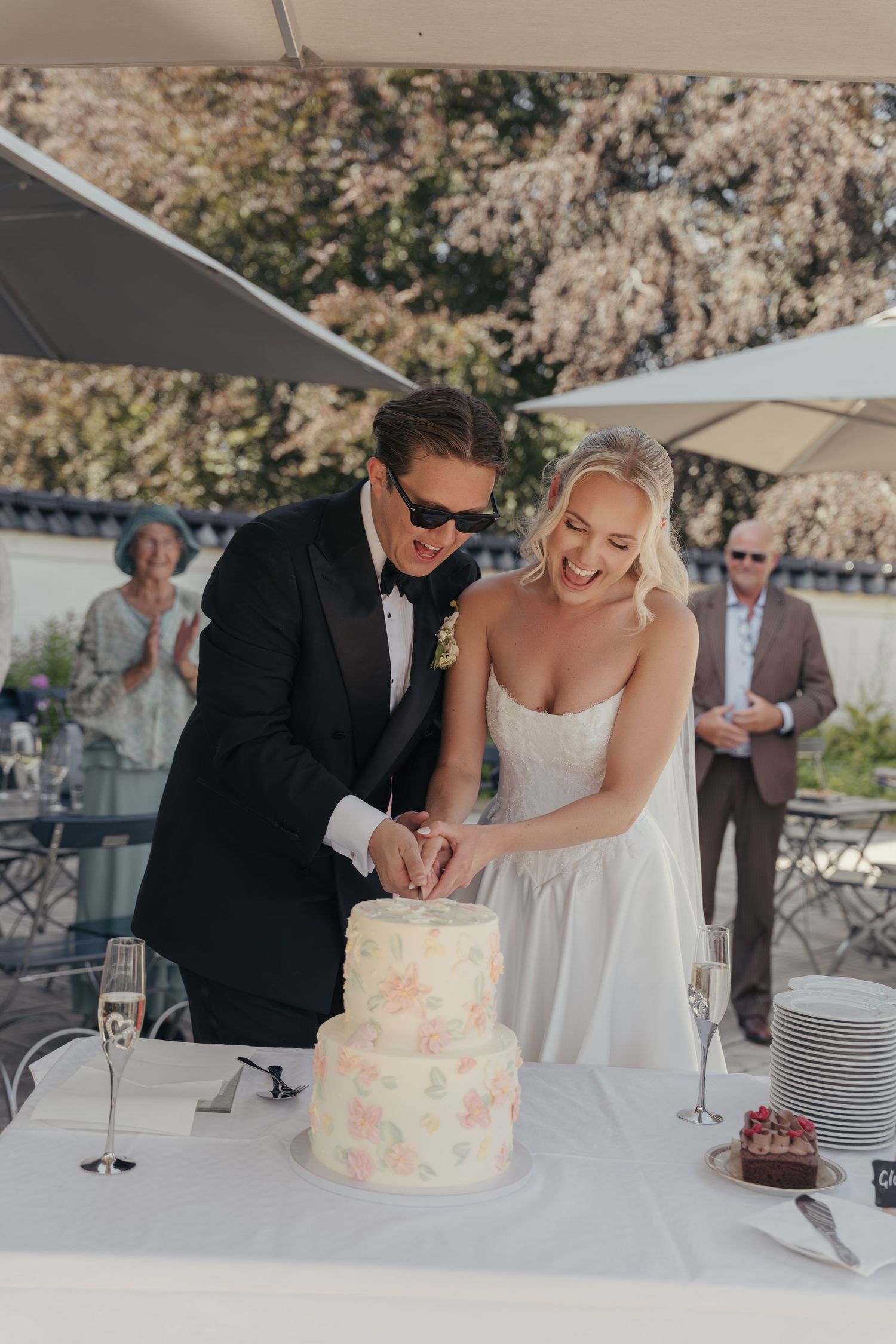 Bride and groom laugh together while cutting a pink floral wedding cake at an outdoor reception with guests watching.