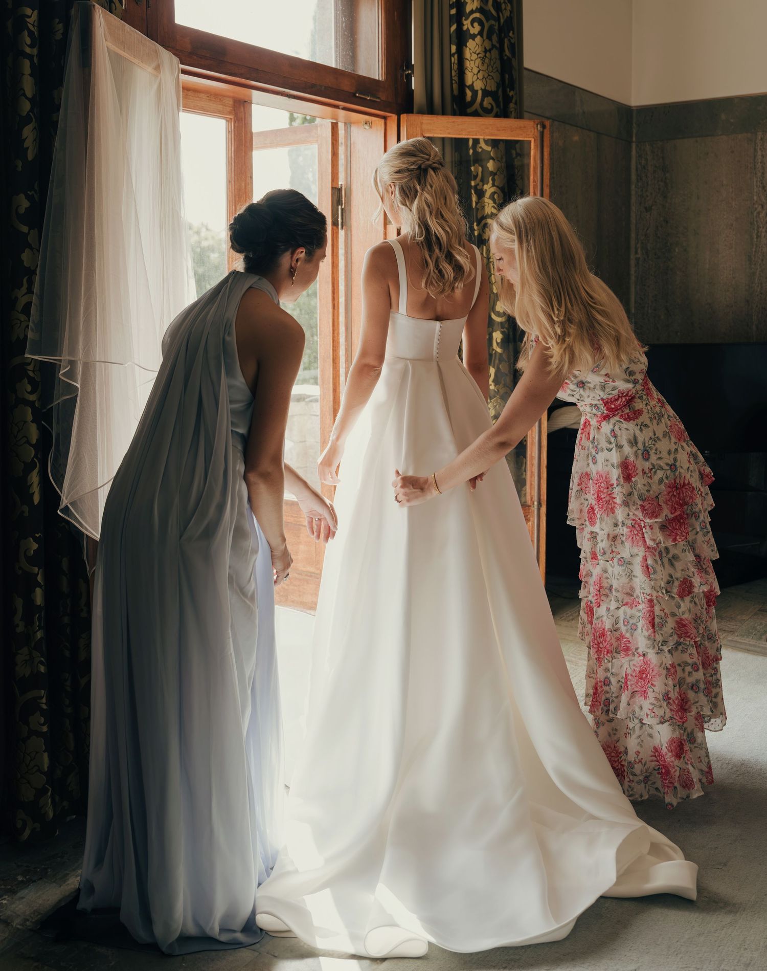 Two bridesmaids in a gray gown and floral dress help a bride in a white wedding dress with a long train by a sunlit window.