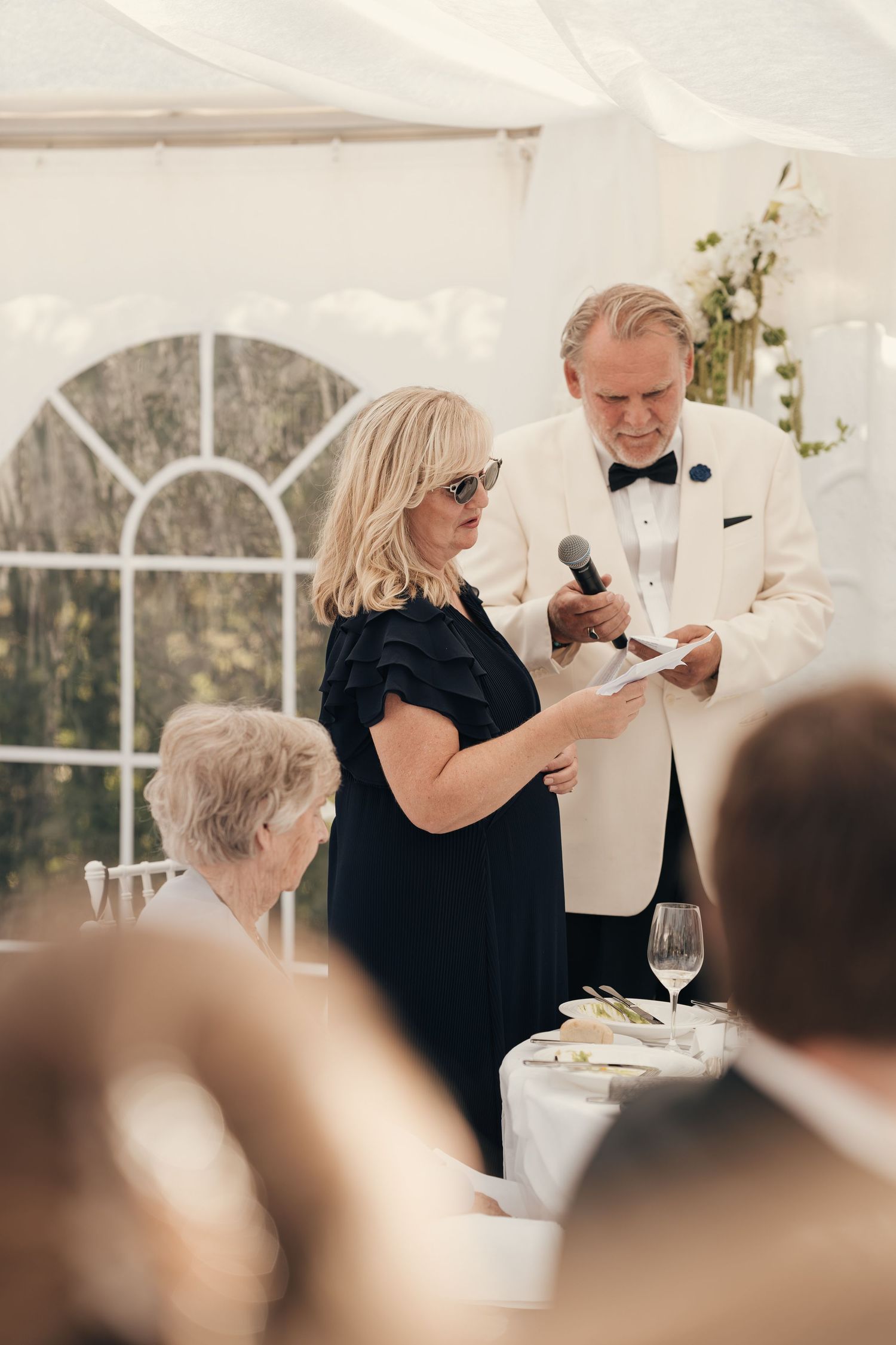 Woman in a navy dress and sunglasses reads a speech while a man in a white tuxedo jacket holds the microphone at a tented wedding reception.
