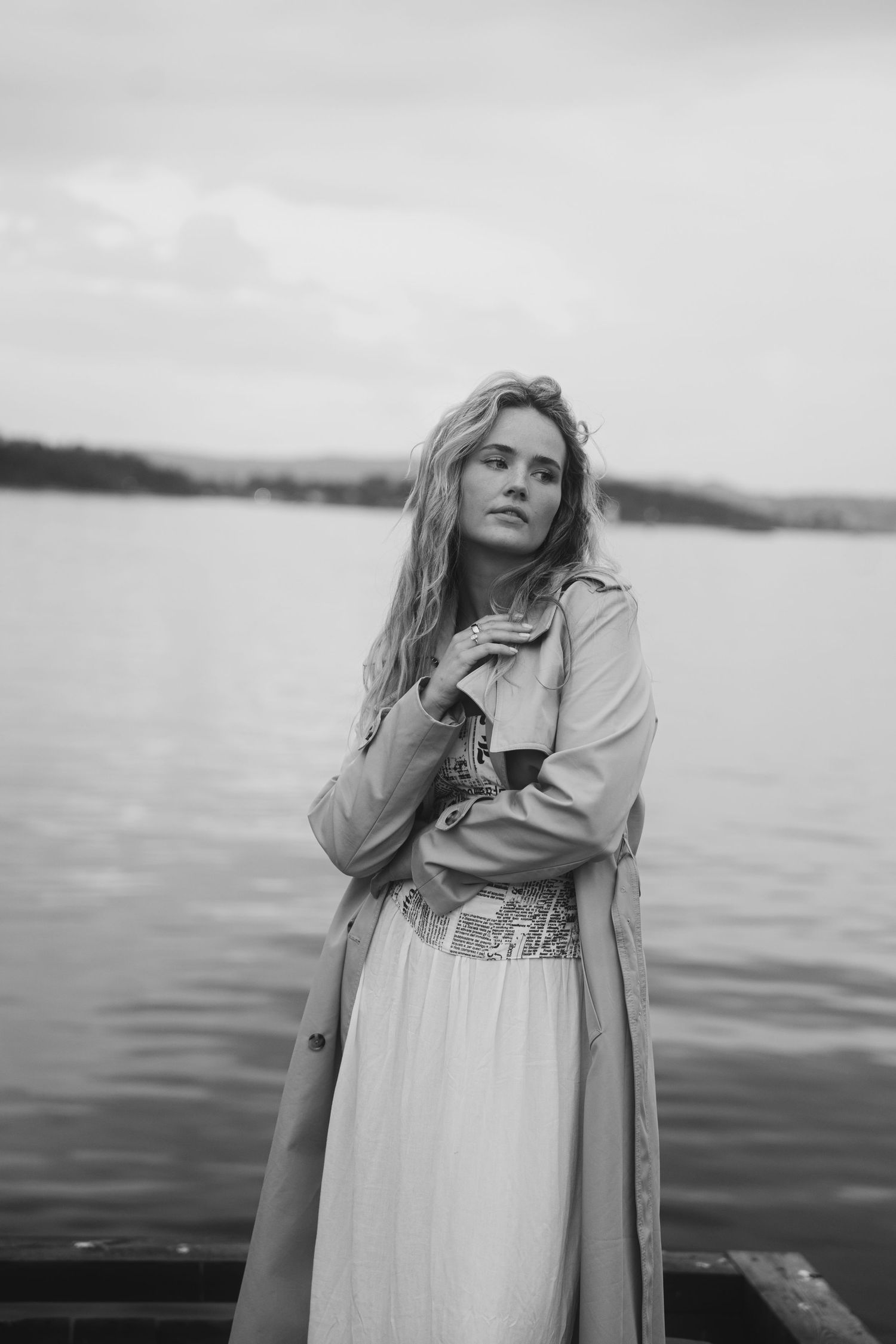 A series of black and white photos showing a woman in a white dress and coat posing near a serene waterfront.