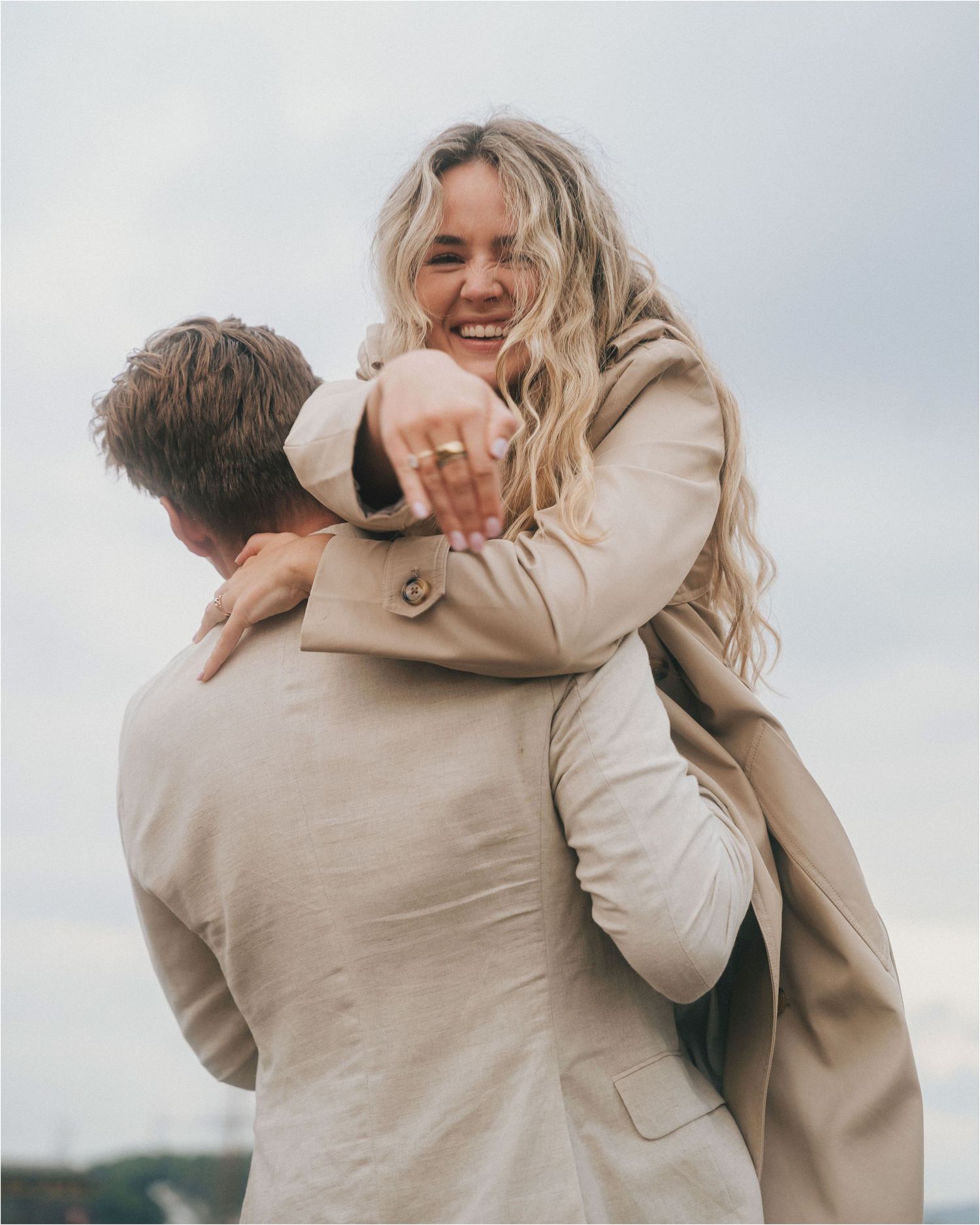 A joyful couple wearing beige coats embrace outdoors with one person being carried piggyback style against a cloudy sky.