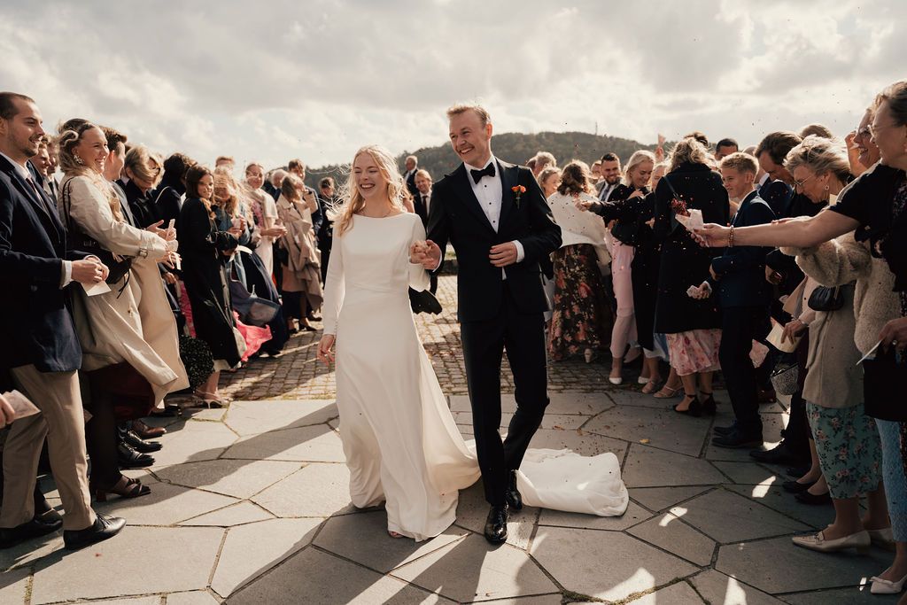 A couple walks down a stone path between rows of guests during an outdoor wedding ceremony on a cloudy day.