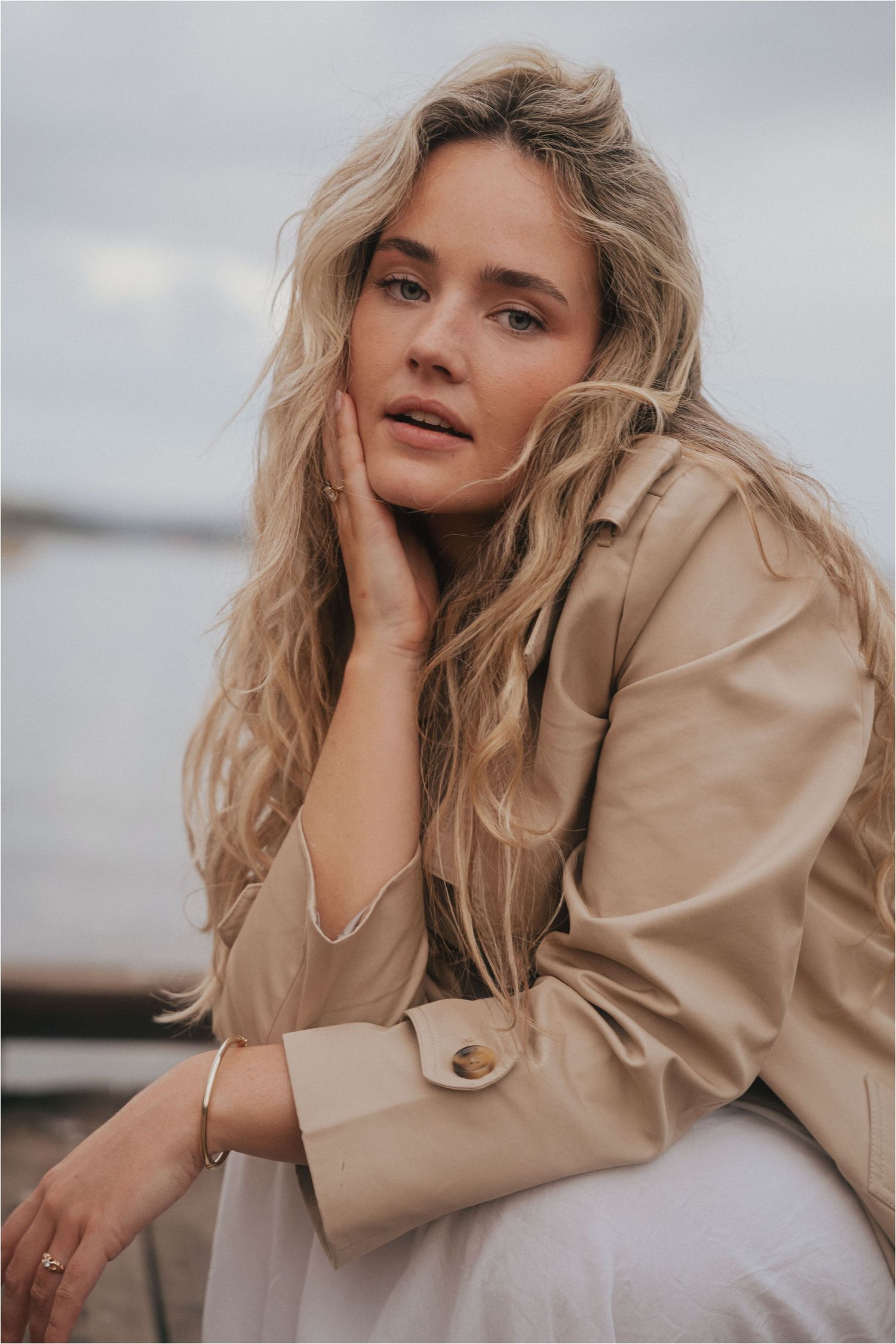 A woman in a beige trench coat poses against a neutral background in a series of soft, moody portrait shots.