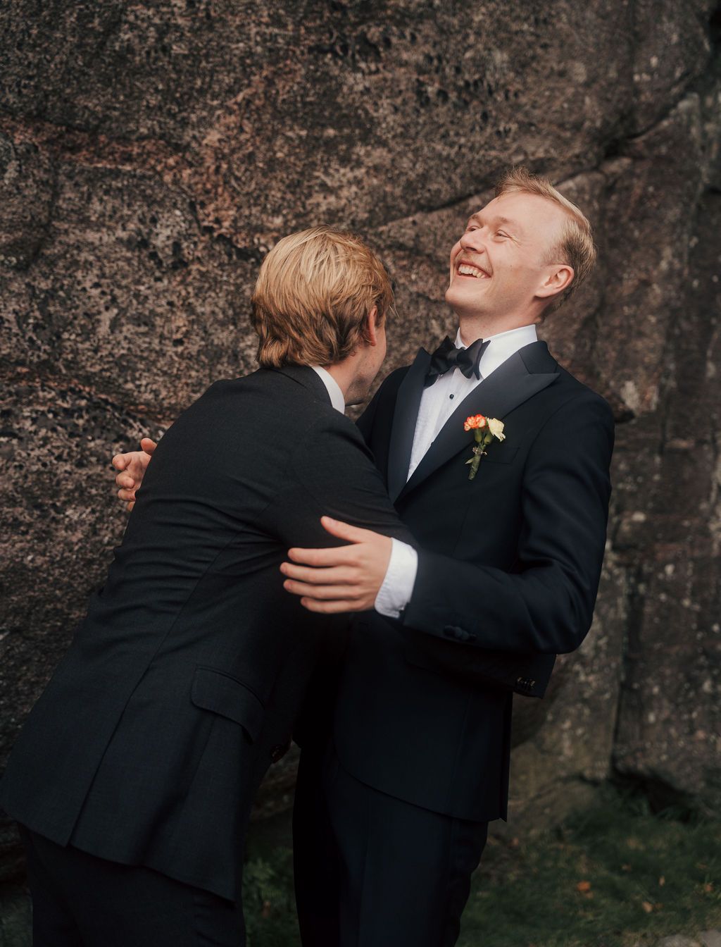 Two people in formal black suits share a candid moment of laughter and embrace against a dark textured wall.