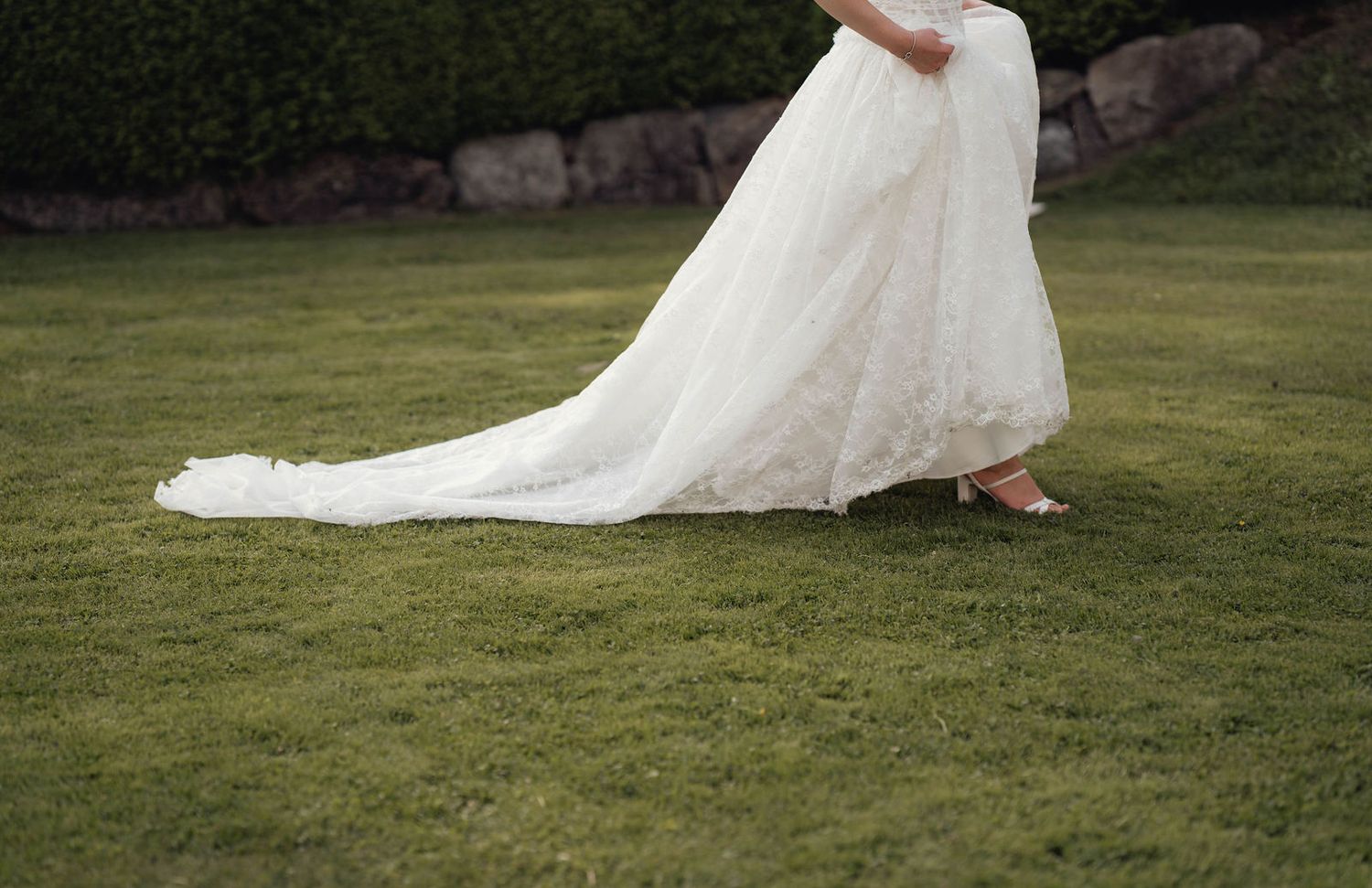 Bride in elegant white lace wedding gown with long train walking on green grass outdoors.