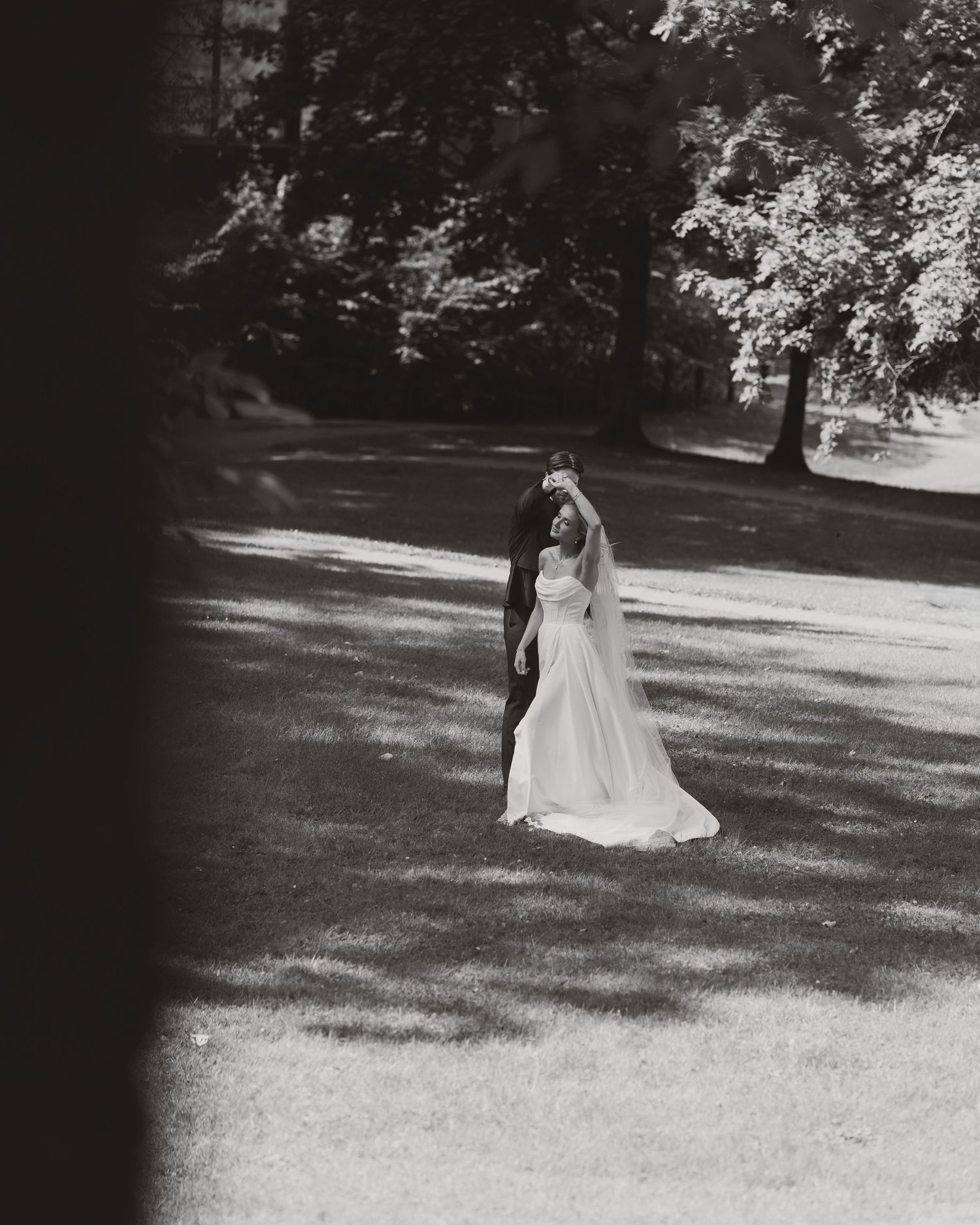 Black and white photo of a bride in a white gown and veil standing gracefully in a sunlit garden.