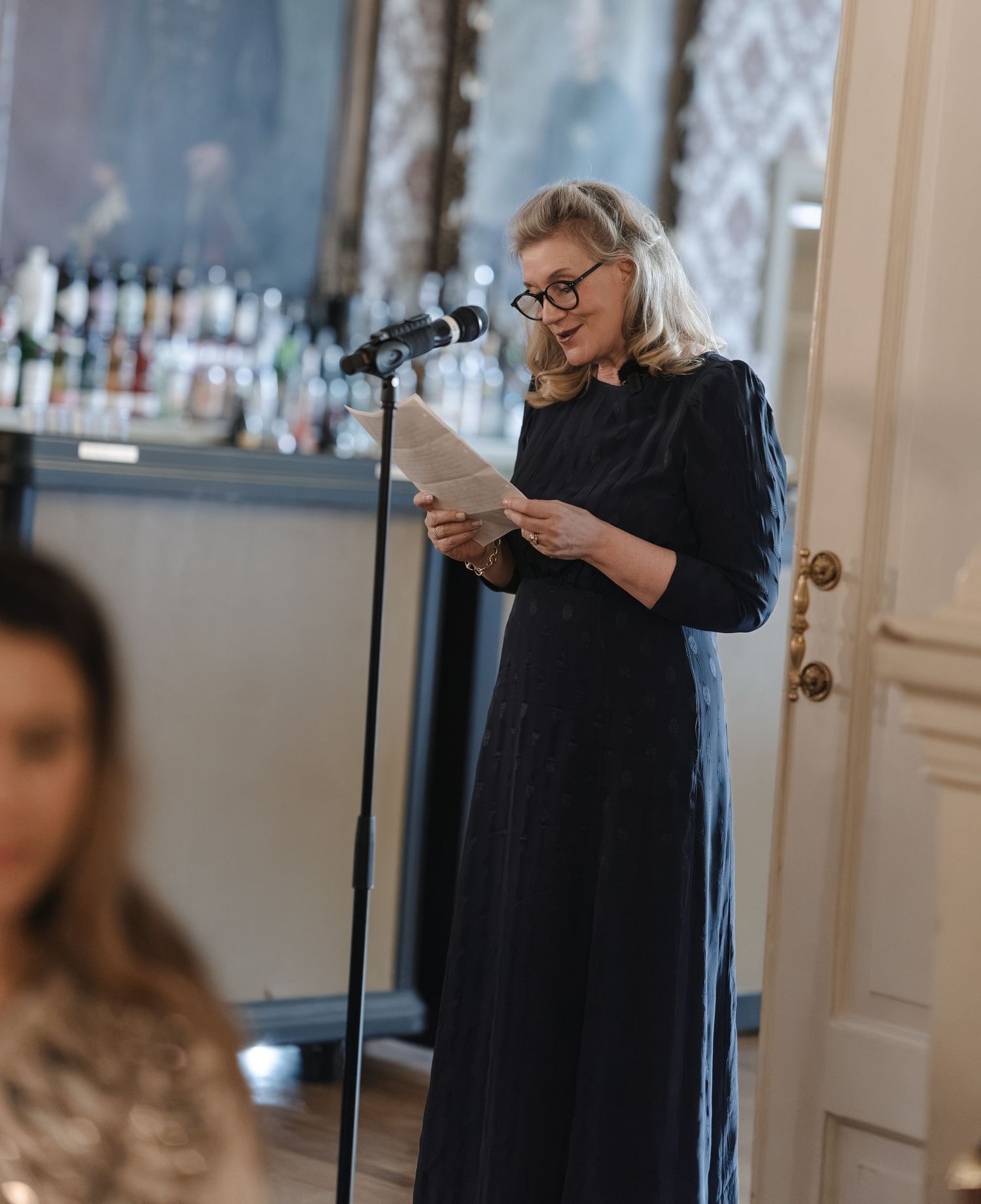 Woman in glasses and a long dark dress reads from papers at a microphone in an elegant room with a bar behind her.