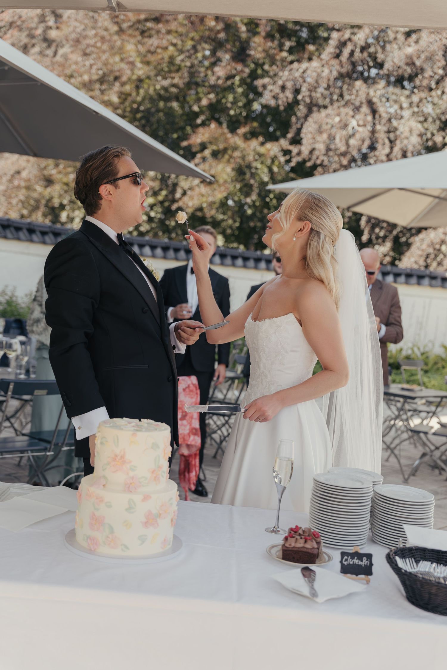 Laughing bride in a white gown and veil feeds a bite of wedding cake to the groom at a sunny garden reception.