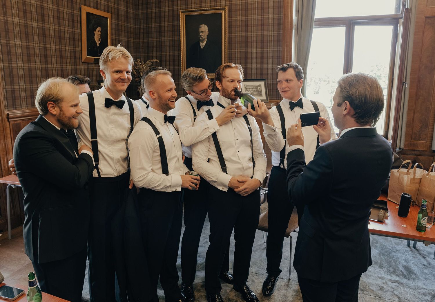 Groomsmen in white shirts, black suspenders and bow ties pose for a photo in a plaid-walled room with framed portraits.