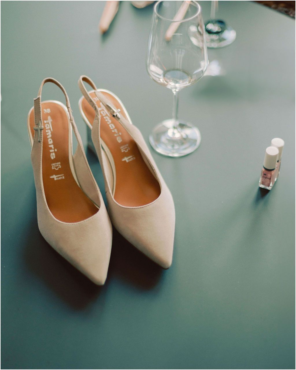 Elegant beige slingback heels with pointed toes displayed against a teal background with wine glasses in soft focus.