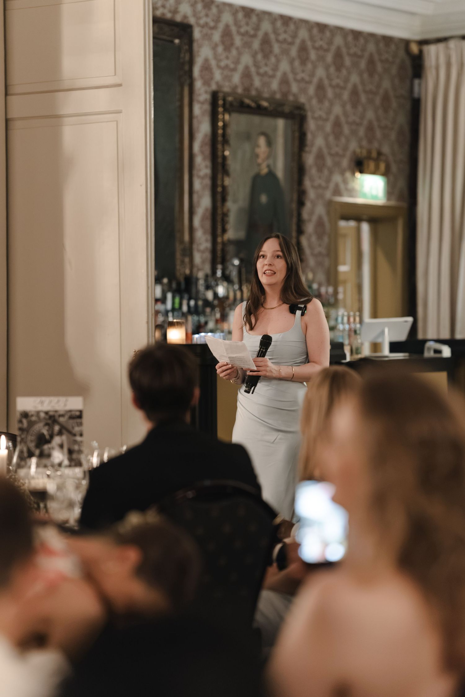 Woman in a gray dress holds a microphone and notes while giving a speech to seated guests in an elegant room.