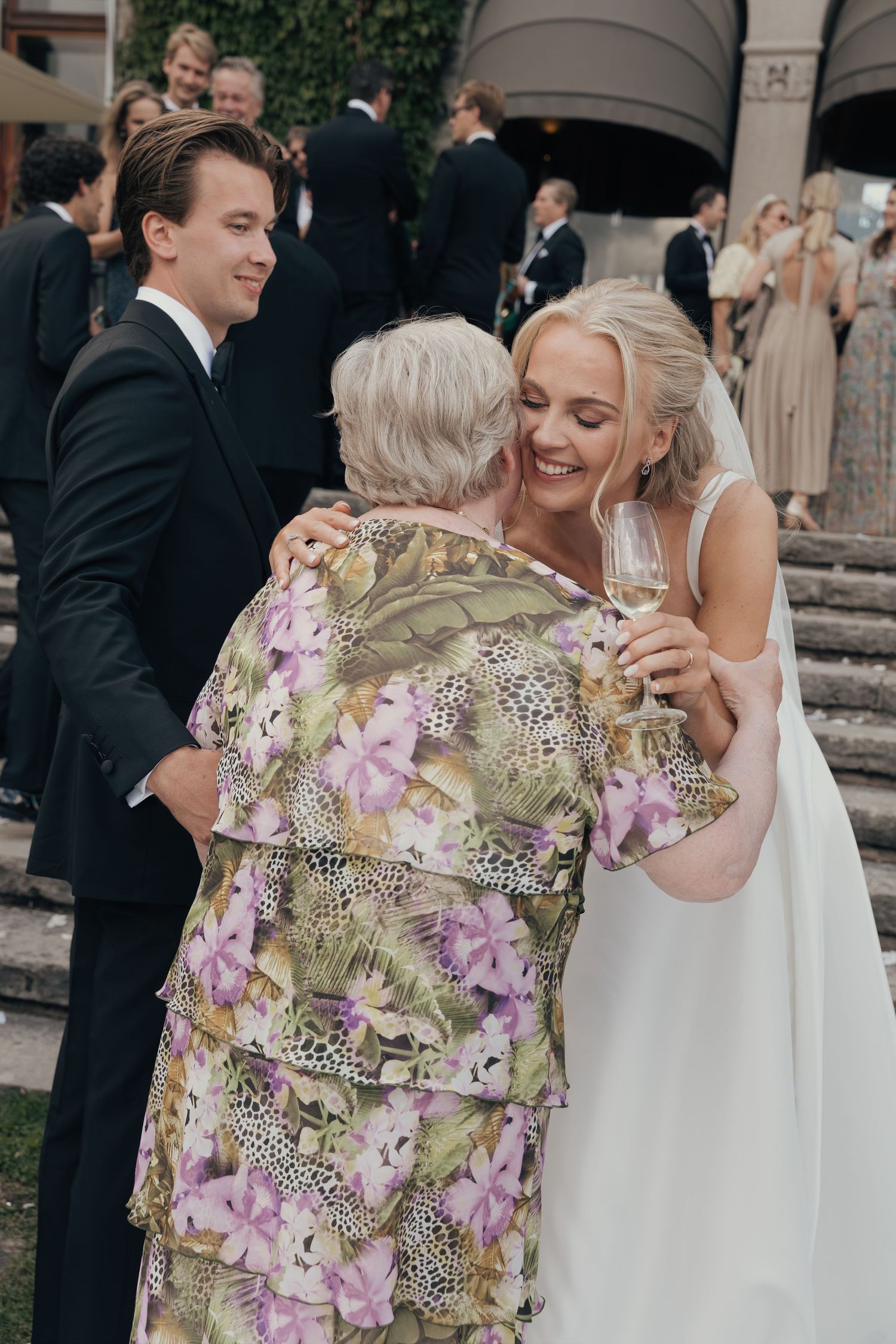 A smiling bride in a white gown hugs an older woman in a floral dress while wedding guests gather on stone steps.