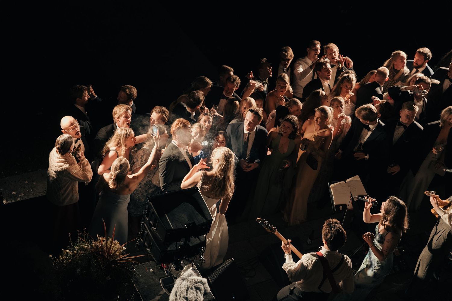 Wedding guests gather closely on a dark stage, singing and playing instruments together in a warmly lit evening performance.